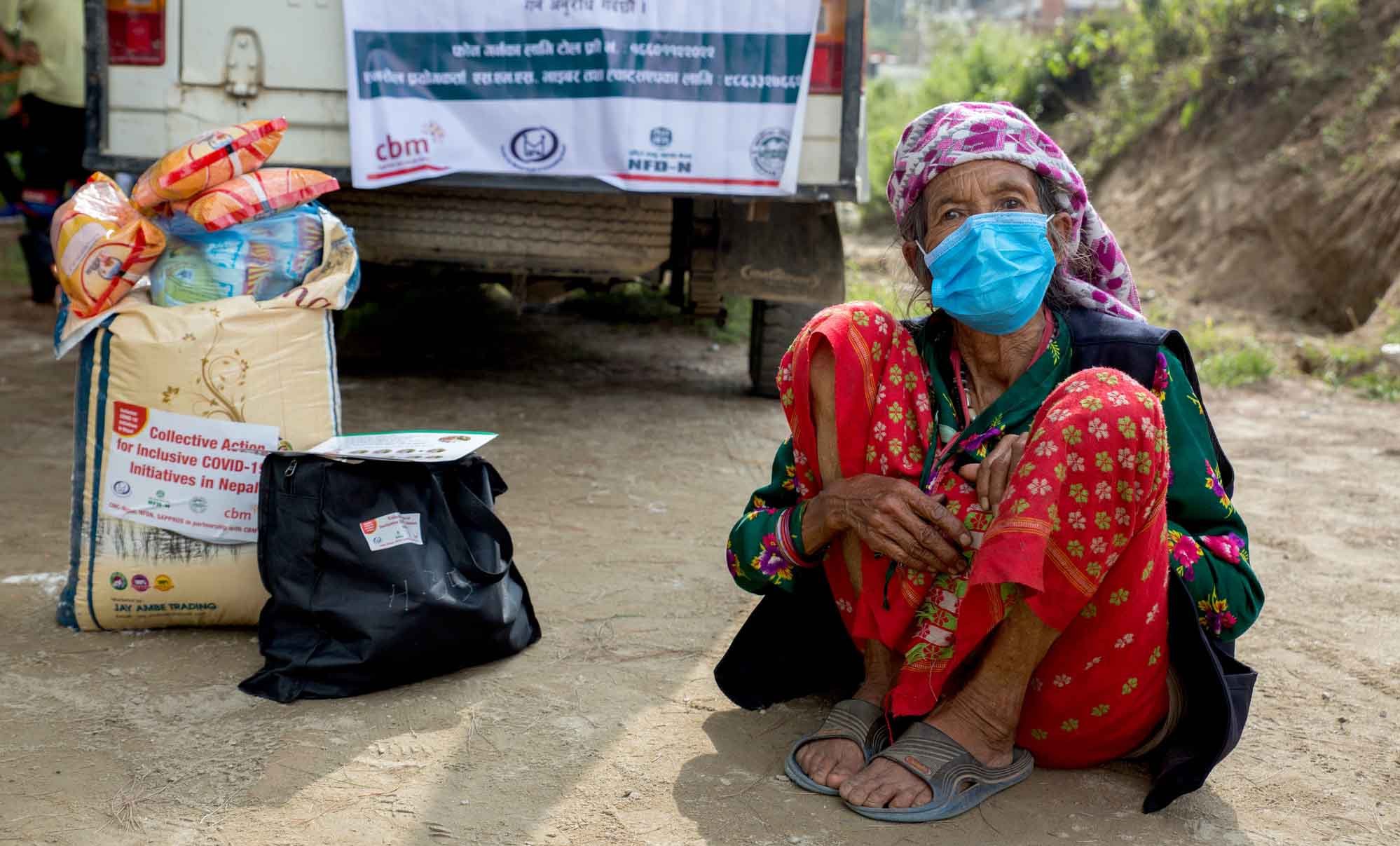 Eine alte Frau aus Nepal mit Maske sitzt in der Hocke. Neben ihr ein Sack Reis oder Mehl sowie weitere Nahrungsmittel.