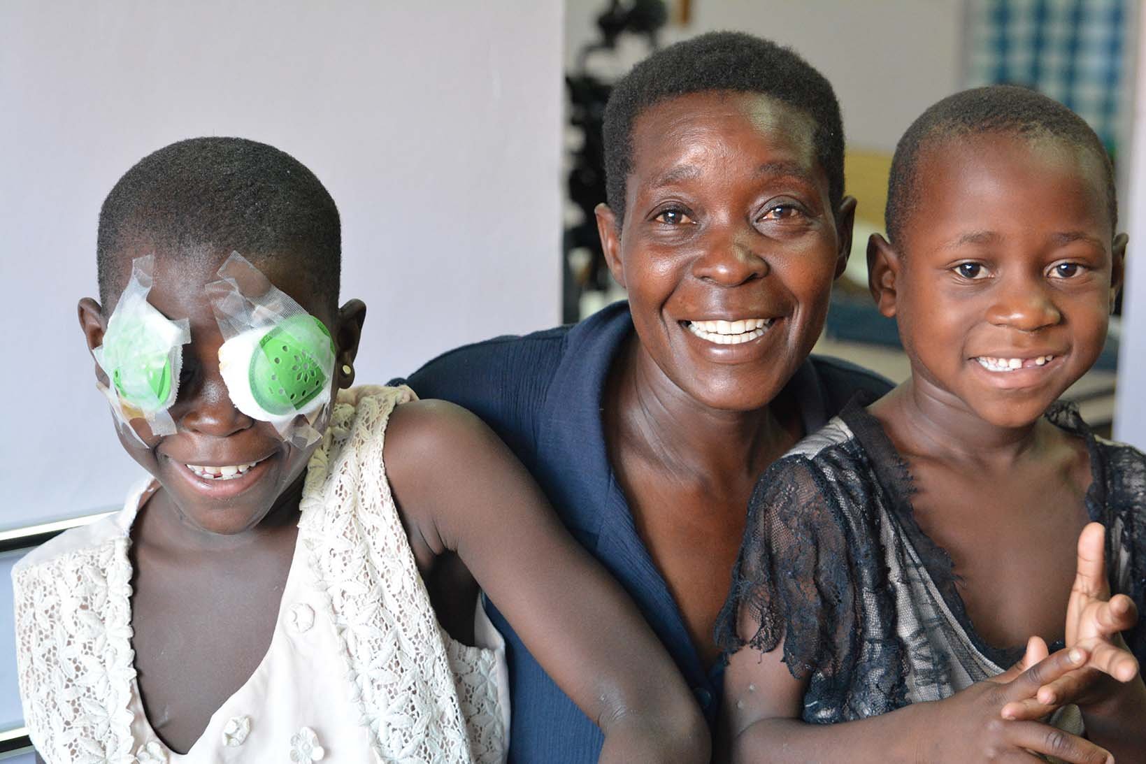 Ein Mädchen aus Uganda mit Augenverband sitzt neben ihrer Mutter und ihrer Nichte auf einem Spitalbett.