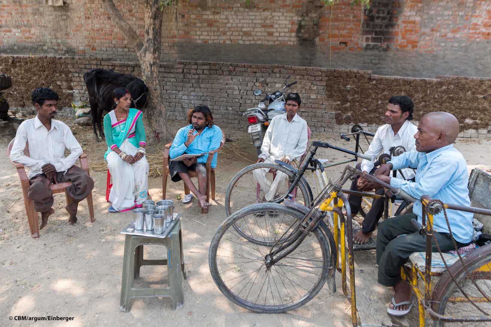 Eine Frau und fünf Männer aus Indien sitzen in einem Halbkreis. Einige von ihnen haben sichtbare körperliche Behinderungen, zwei von ihnen sitzen auf einem Tricycle.