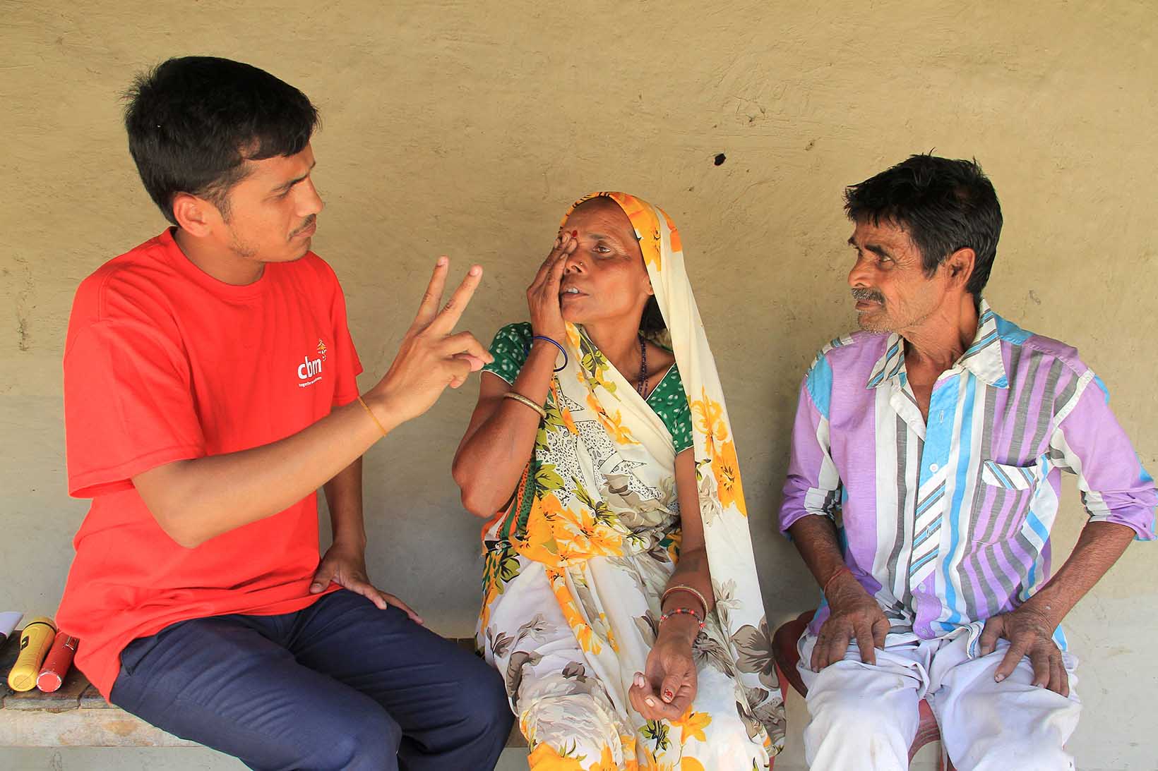 Ein Mann überprüft mit einem Fingerzähltest die Sehkraft einer Frau aus Indien. Die Frau hält mit ihrer rechten Hand ihr rechtes Auge zu, ihr Mann sitzt neben ihr.