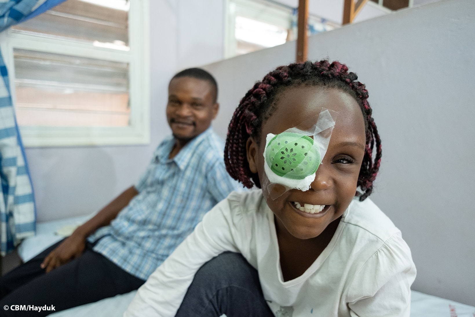 Ein Mädchen aus Uganda trägt einen Augenverband und lächelt, ihr Vater im Hintergrund lächelt ebenso.