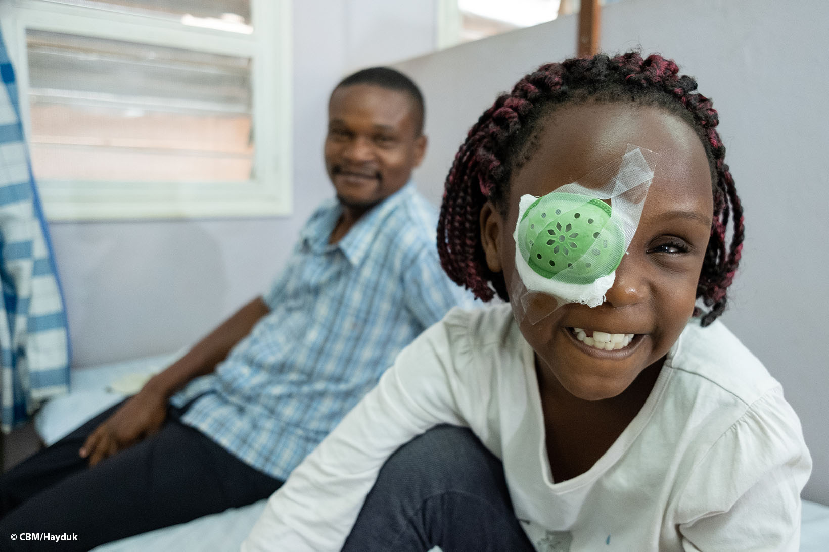 Ein Mädchen aus Uganda, das einen Augenverband trägt, lächelt in die Kamera. Hinter ihr sitzt ihr Vater, der ebenfalls lächelt.