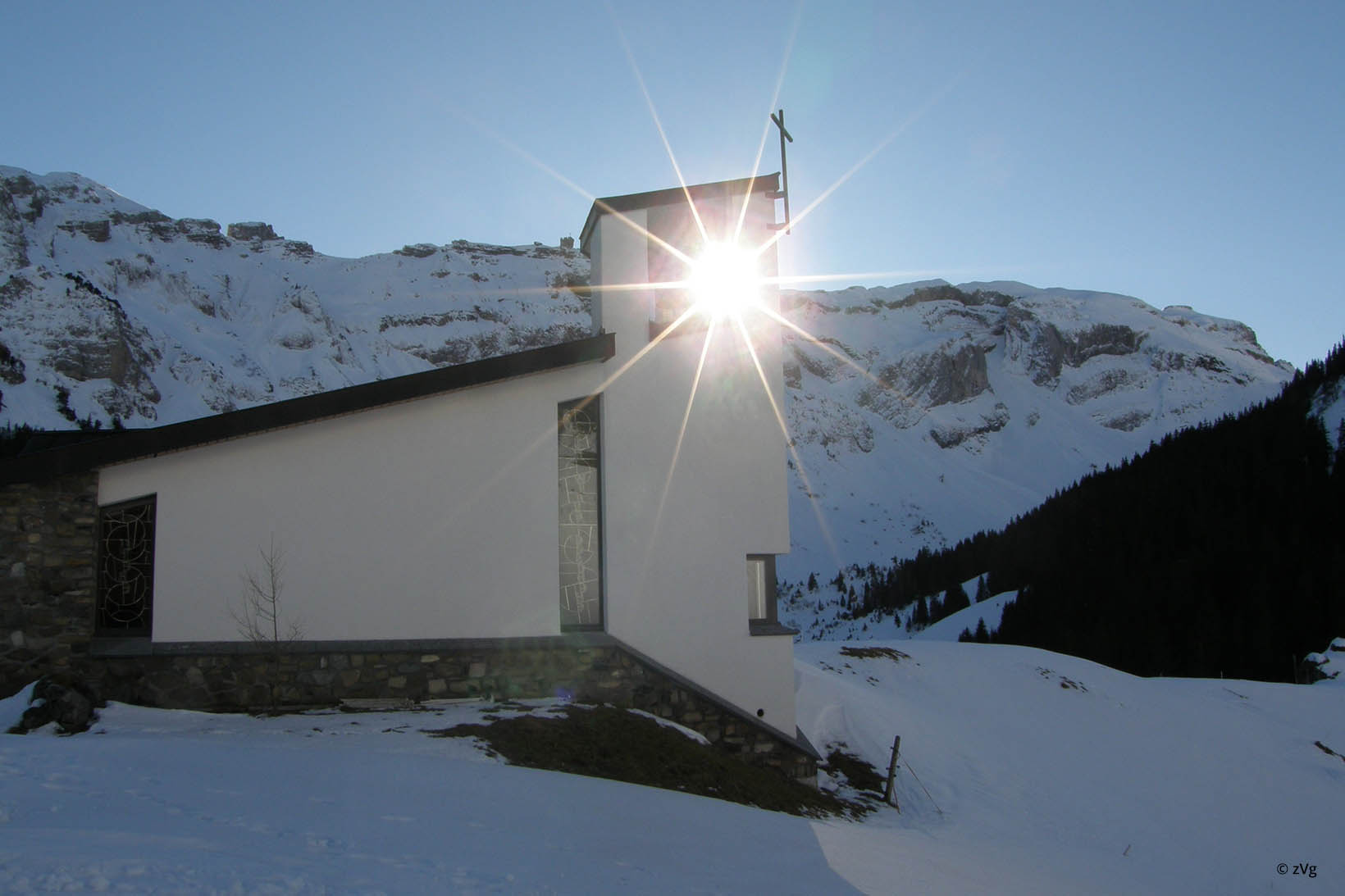 Le soleil se réfléchissant sur la fenêtre d’une église. Celle-ci est entourée d’un paysage montagneux sous la neige.
