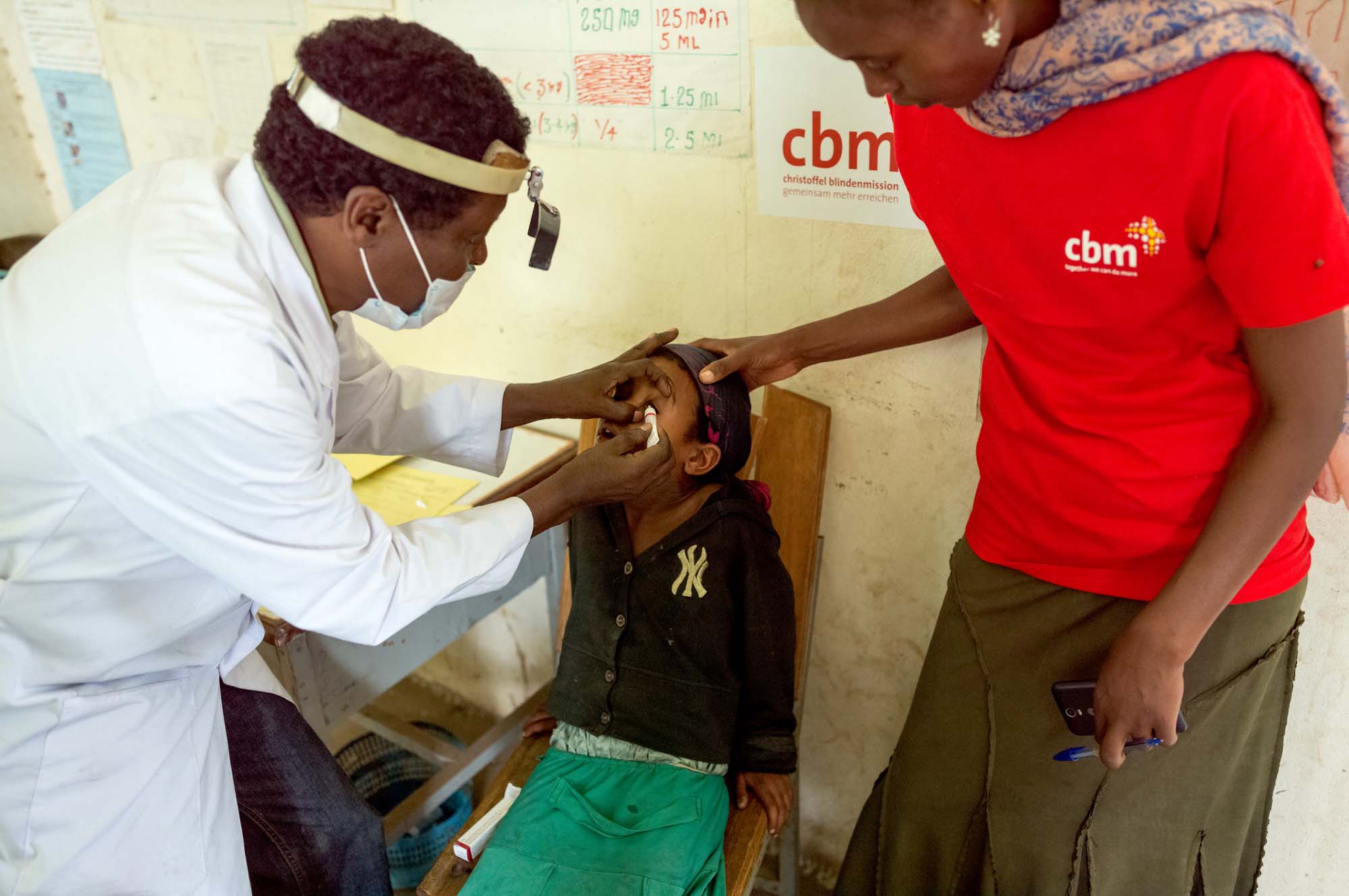 Ein Augenarzt salbt das Auge eines Mädchens ein. Neben dem Mädchen steht eine CBM-Helferin.
