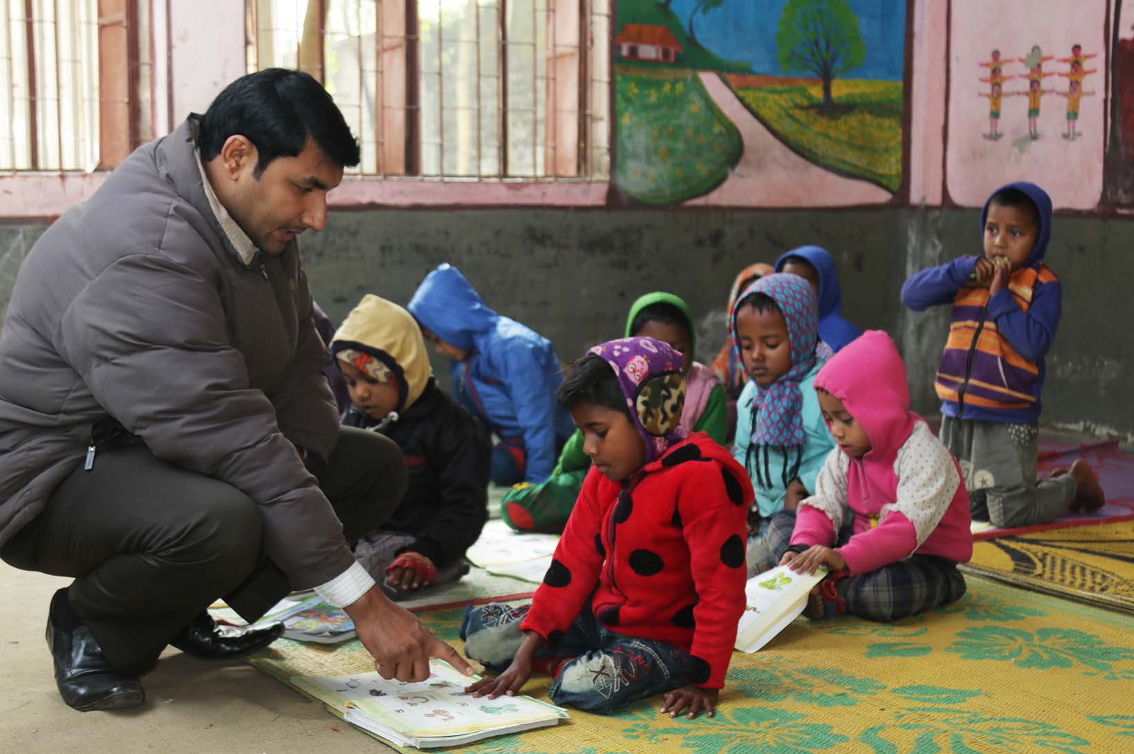 Kinder mit Jacken und Mützen sitzen auf dem Boden vor ihren Schulbüchern. Der Lehrer ermutigt Ripon, zu sprechen.