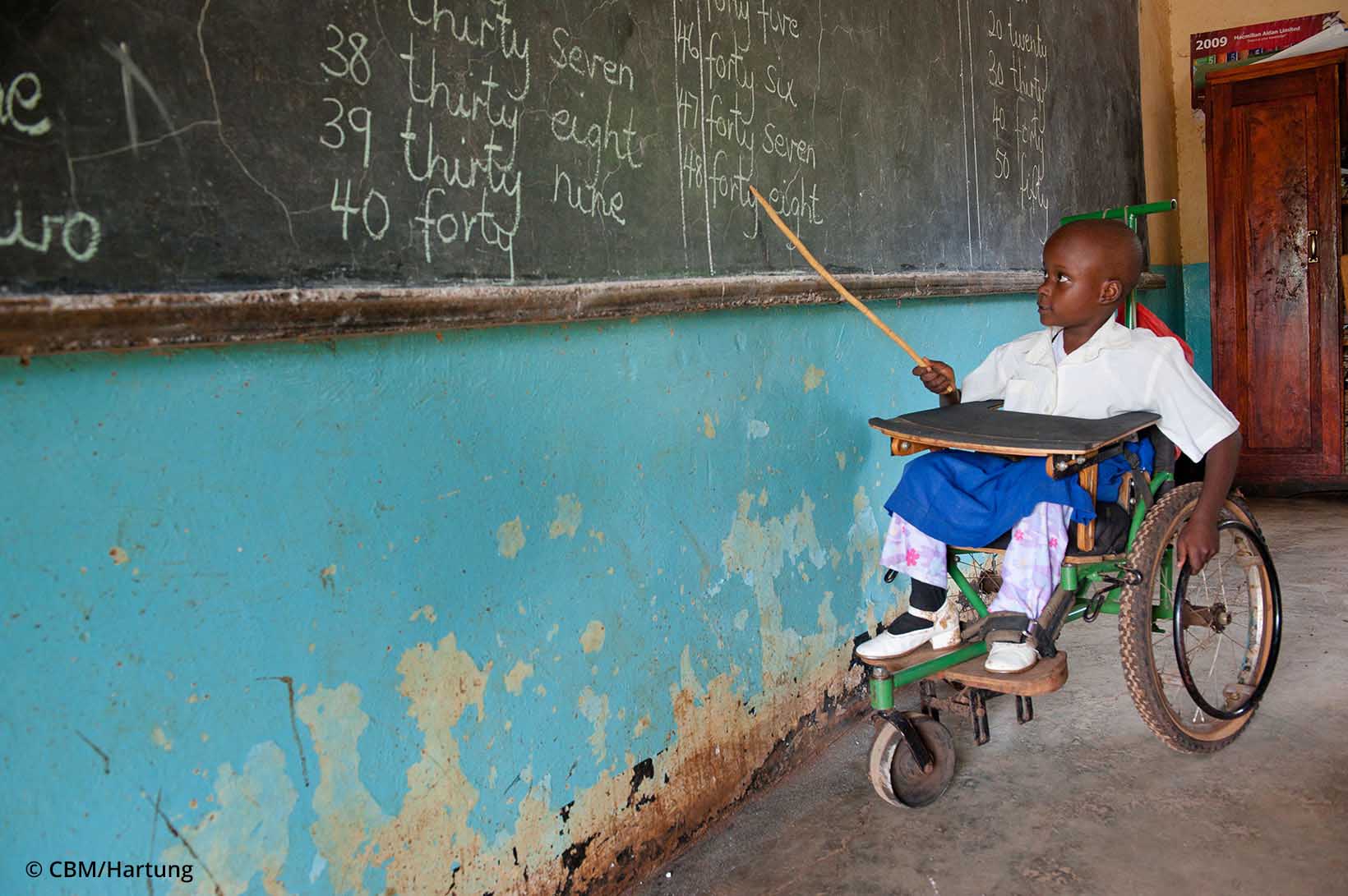 Une fillette de Tanzanie en chaise roulante pointe une canne sur le tableau à l'école.