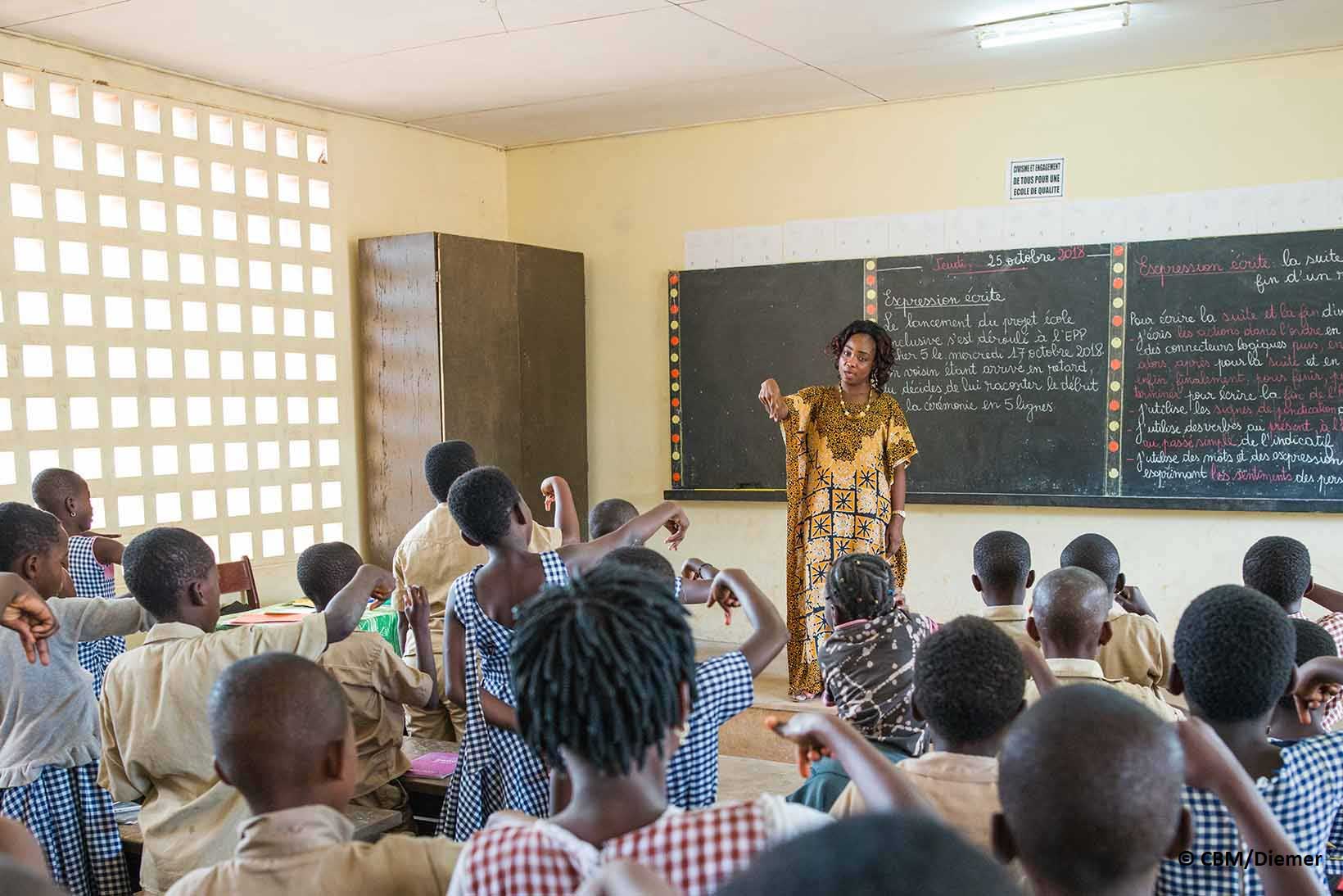 Eine Lehrerin zeigt vor der Klasse Ausdrücke in Gebärdensprache, alle Kinder üben sie.