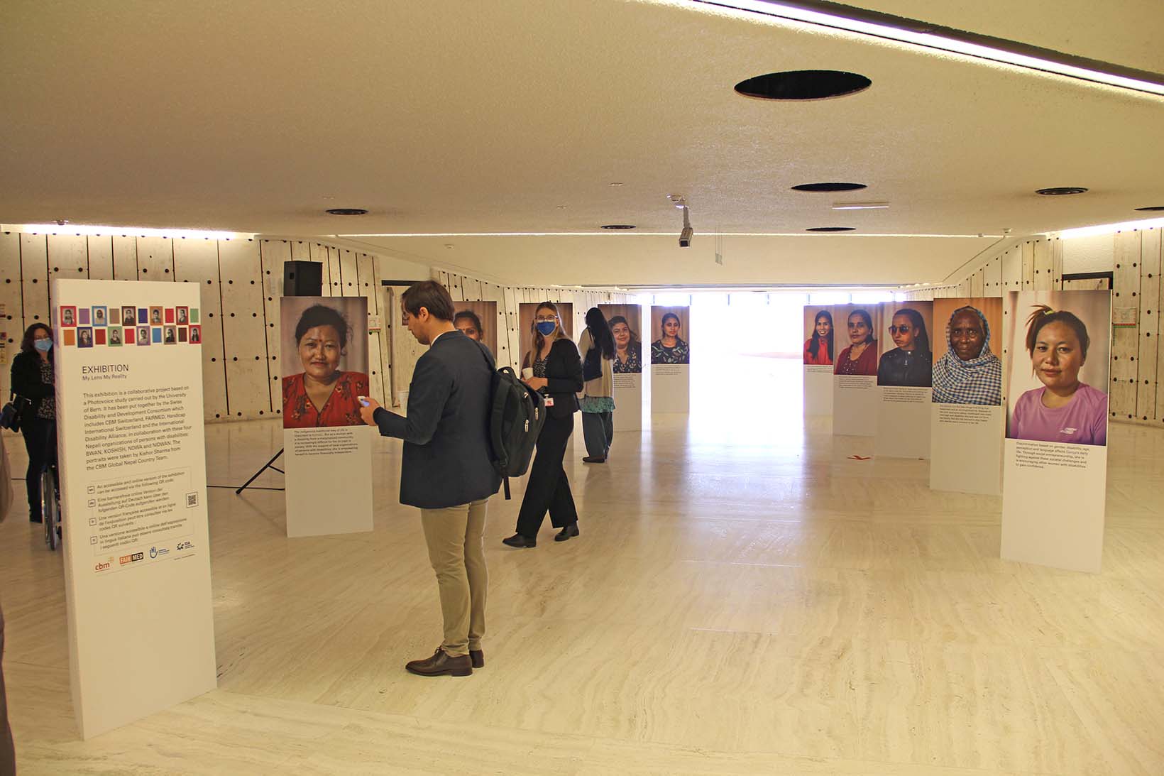 Eine Fotoaustellung in einem Gebäude der UNO in Genf. Links vorne ist ein Panel, das den Kontext der Ausstellung beschreibt, dahinter sowie rechts davon sind zehn Panels mit Portraits von nepalesischen Frauen. Einige Personen schauen sich die Panels an.