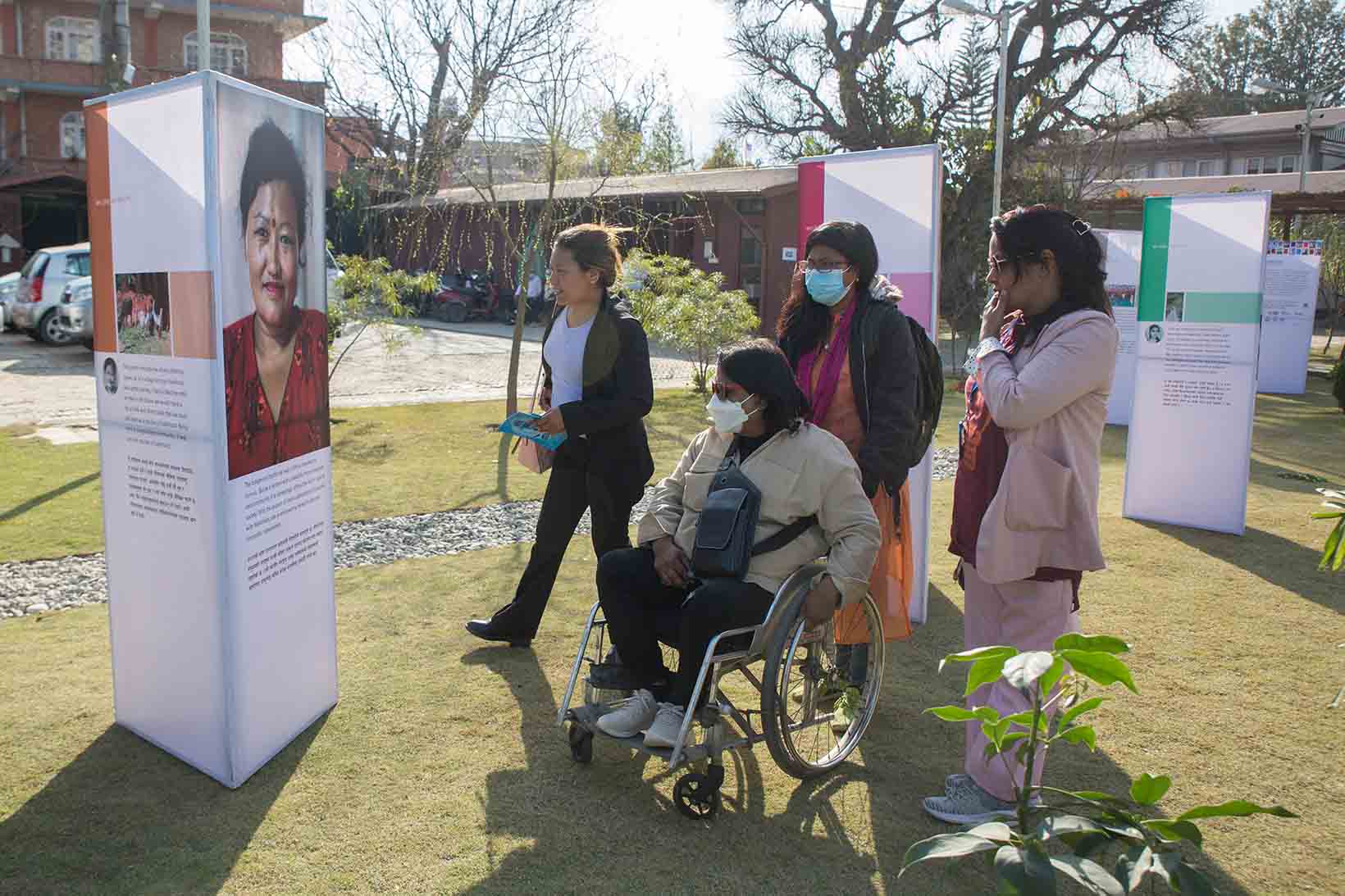 Einige Frauen aus Nepal, eine davon ist im Rollstuhl, schauen sich ein Panel mit dem Porträt einer Frau an.