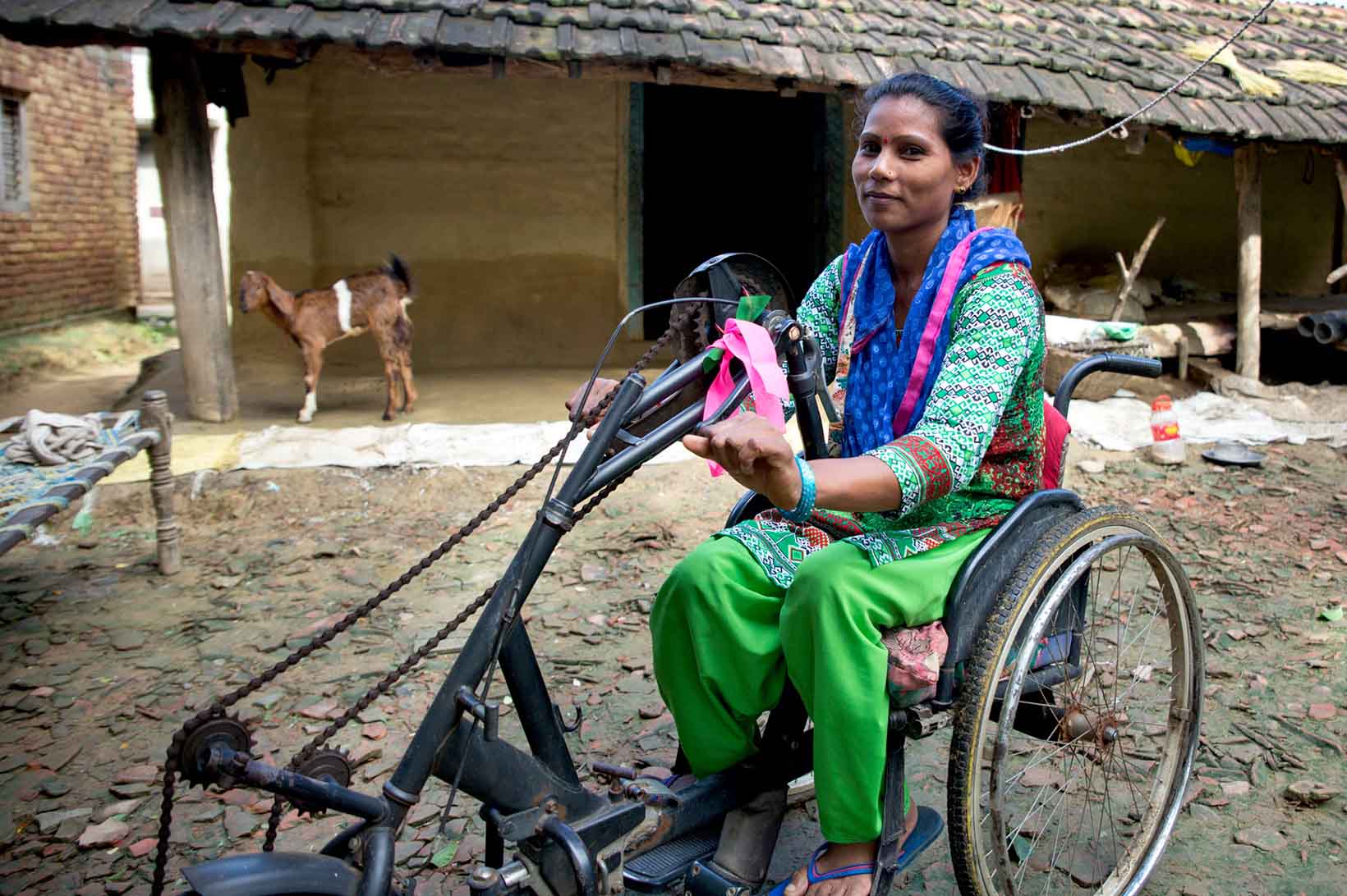Eine junge Frau aus Nepal sitzt in einem Tricycle vor ihrem Haus. Im Hintergrund steht eine Ziege.