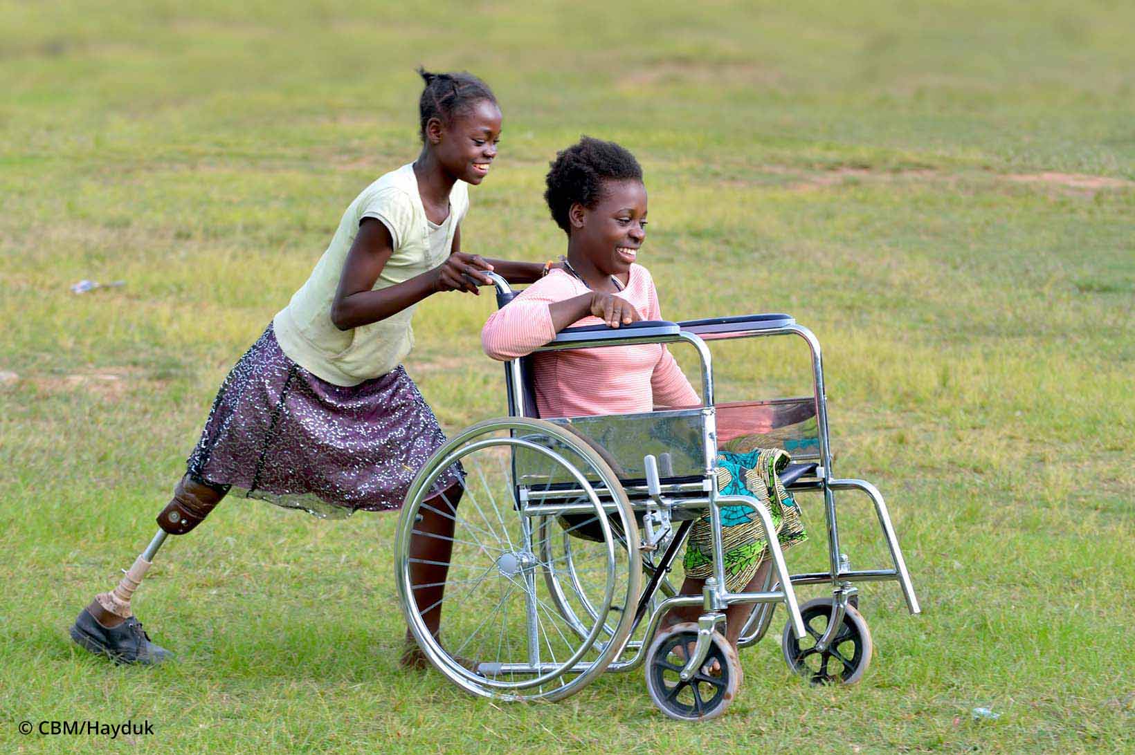 Eine junge Frau mit Beinprothese schiebt die Kollegin an, die im Rollstuhl sitzt.