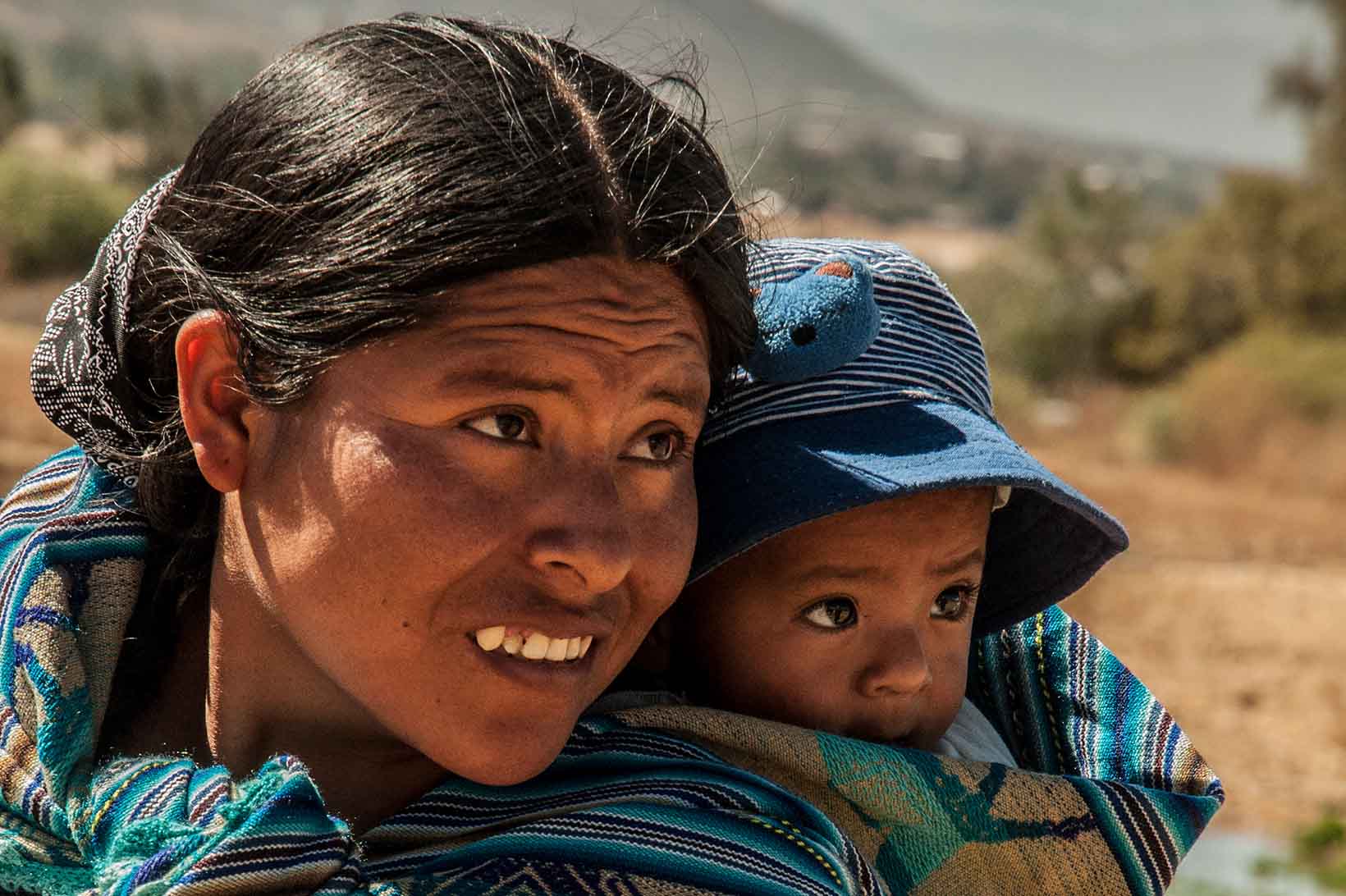 Eine junge indigene Frau aus Bolivien trägt ein Kleinkind in einem Tragetuch auf dem Rücken. Den Kopf haben sie nach links gewendet.