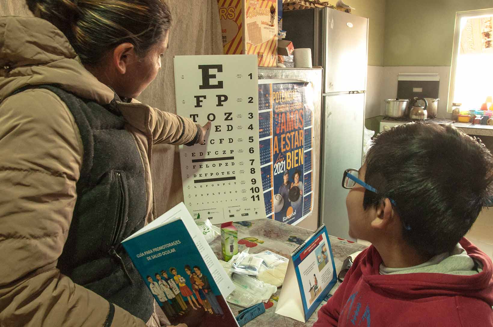 Eine Frau aus Bolivien zeigt mit einer Hand auf eine Sehtesttafel. Ein Junge mit Brille blickt hin. Mit der anderen Hand hält sie eine Infobroschüre. 
