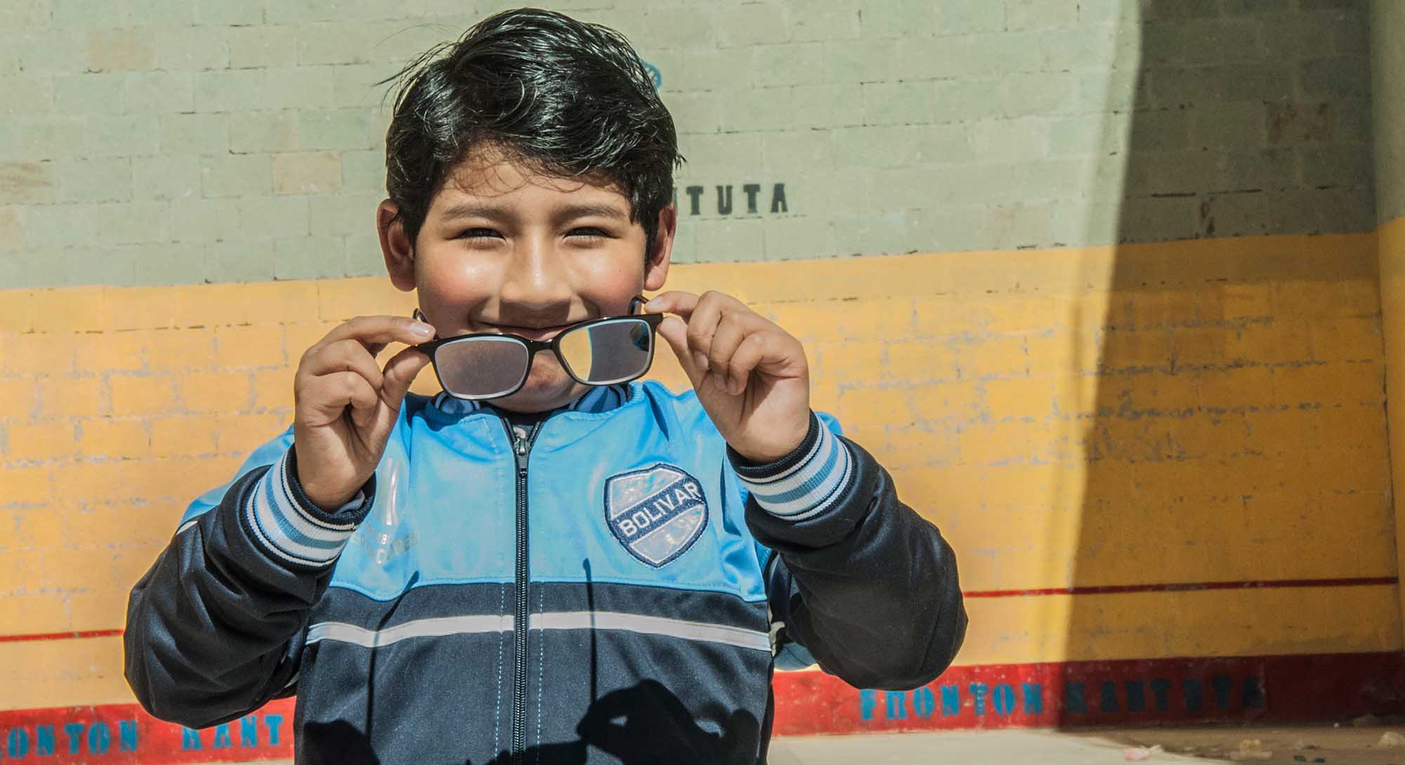 Lächelnd hält ein achtjähriger Junge aus Bolivien seine erste Brille mit beiden Händen gegen die Kamera. 