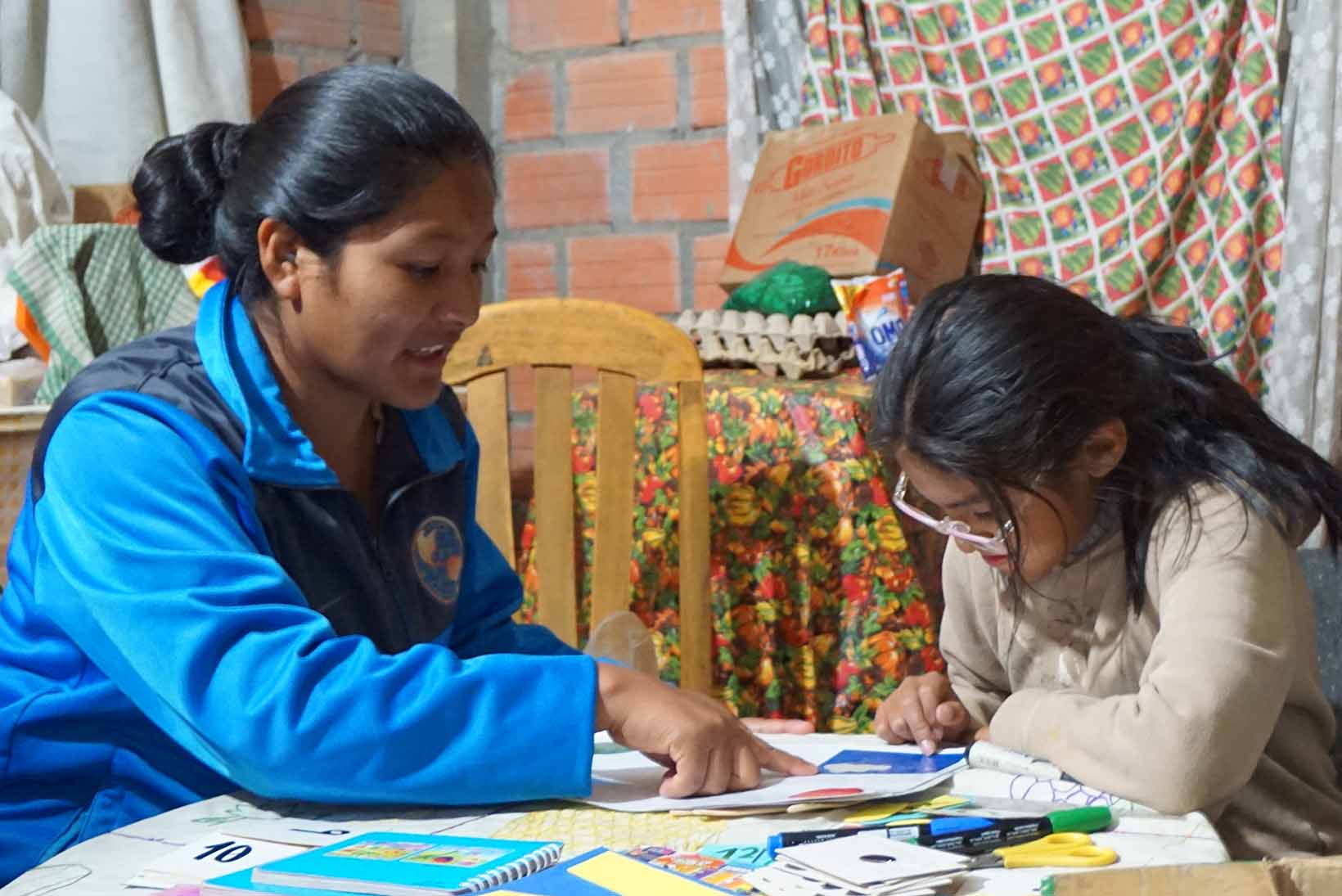 Assise à une table, une fillette bolivienne souffrant d’un déficit d’apprentissage fait ses devoirs en compagnie d’une femme qui lui donne des leçons de soutien.