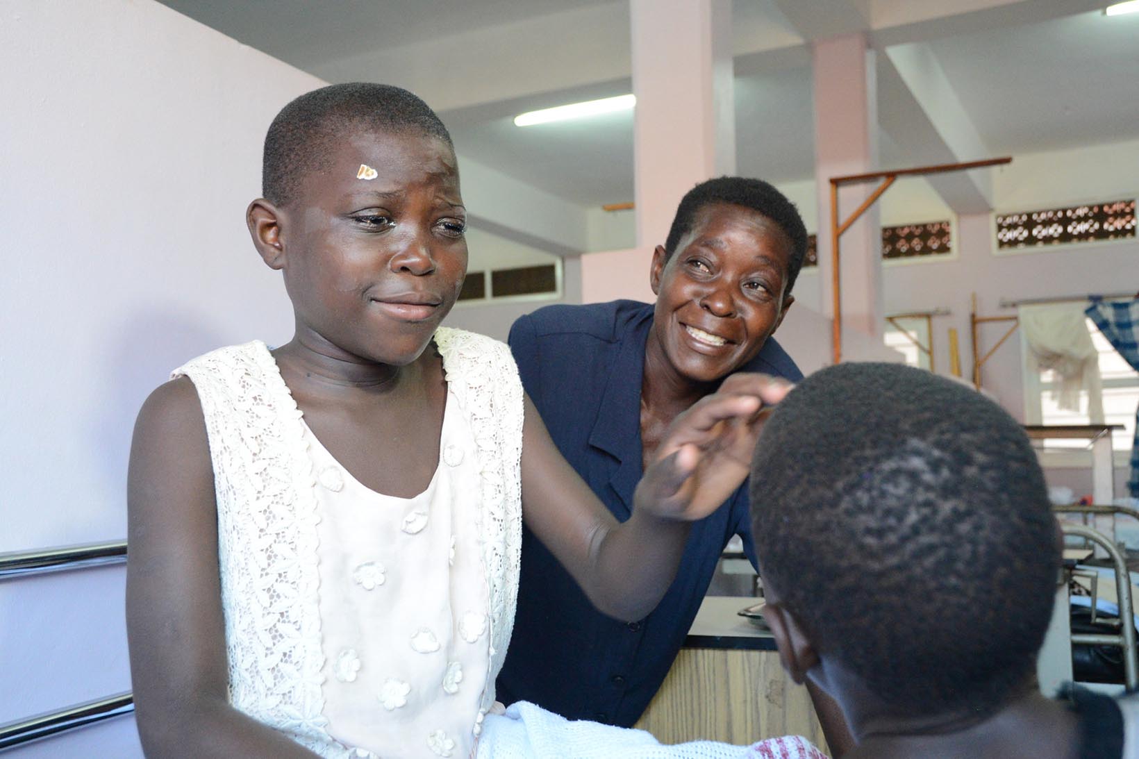 Ein Mädchen aus Uganda, das vor kurzem eine Graue-Star-Operation hatte, blickt gerührt ein anderes Mädchen an. Im Hintergrund lächelt die Mutter des Mädchens, das vor kurzem operiert worden ist.