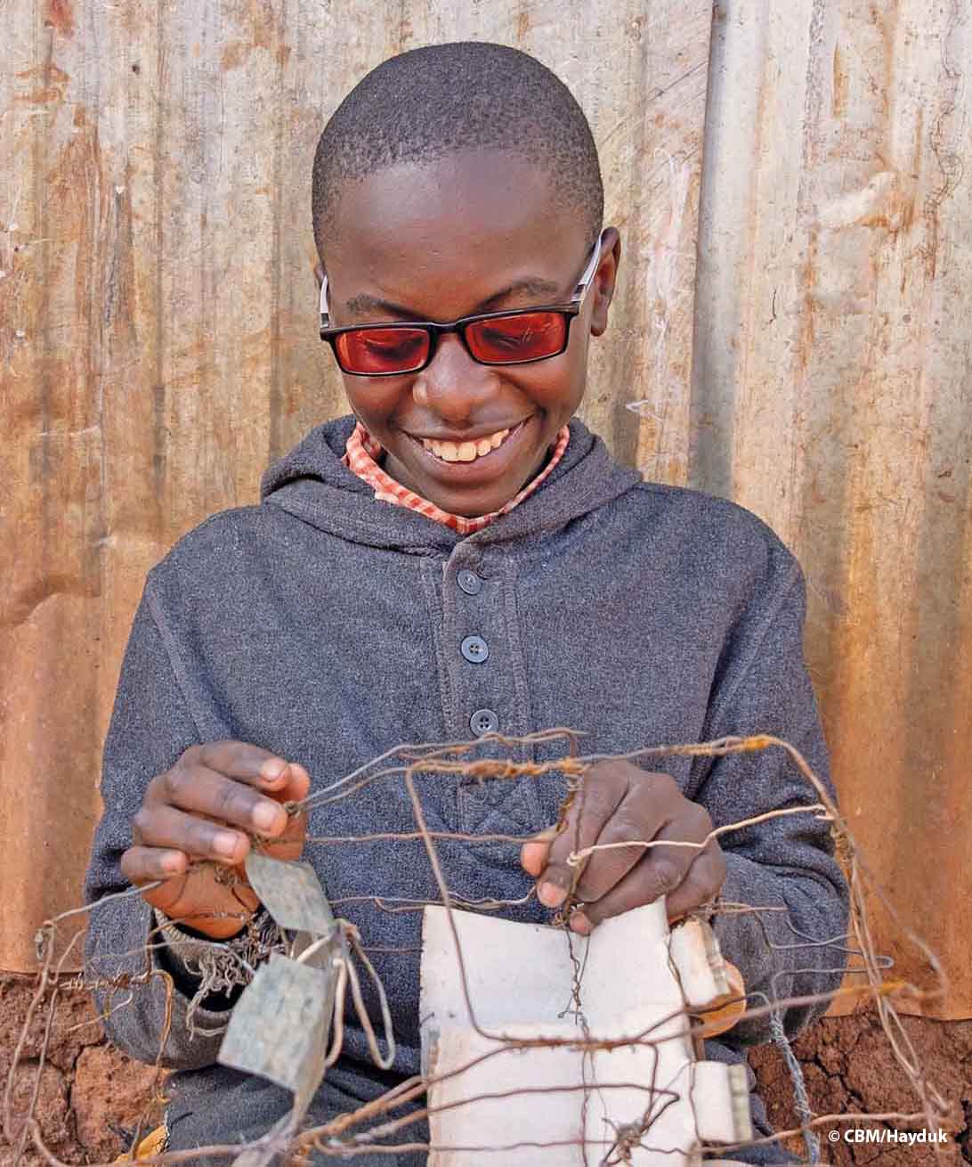 Ein Junge aus Kenia, der eine UV-filternde Sonnenbrille trägt, bastelt auf seinem Schoss ein Auto aus altem Draht mit zwei Sitzen und Rädern aus Verschlüssen von Petflaschen.