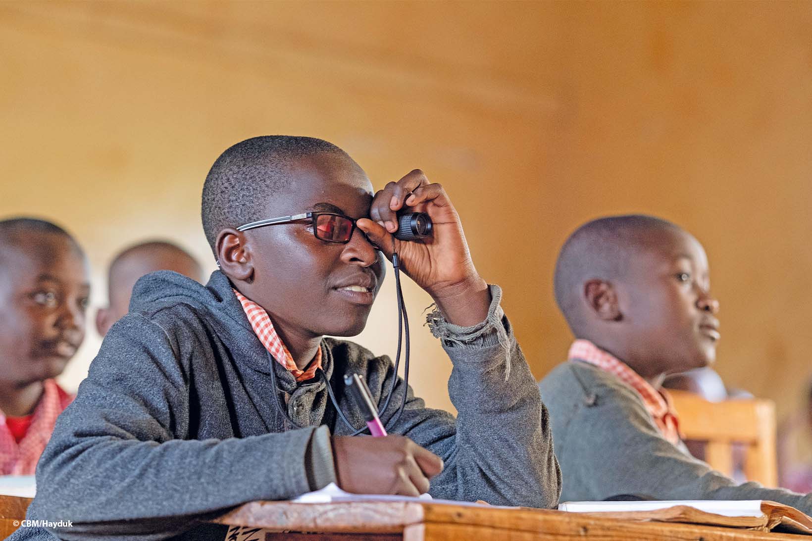 Un jeune Kenyan, qui porte des lunettes de soleil filtrant les UV, regarde le tableau noir avec un télescope tout en souriant. Il porte l’uniforme de son école. 