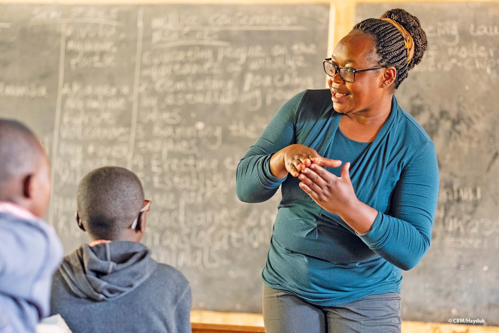 Une institutrice kenyane explique quelque chose à ses élèves. Derrière elle, on aperçoit le tableau noir couvert d’écriture. 