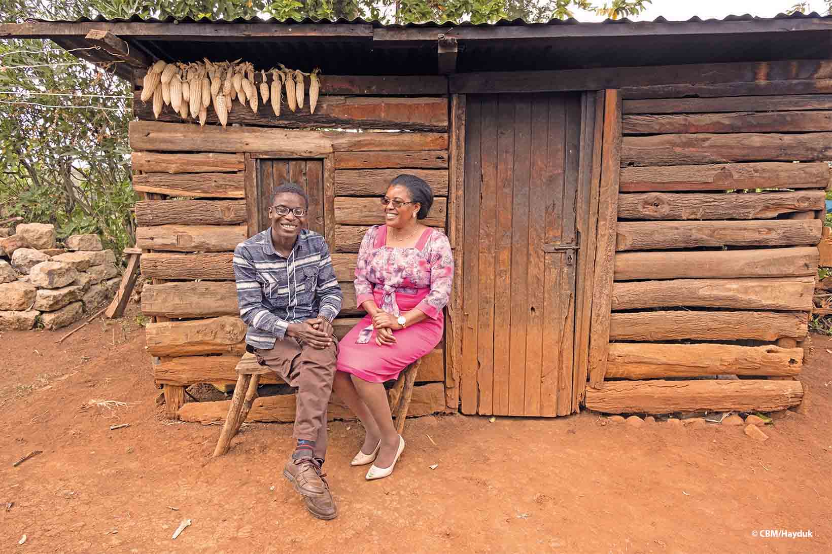 Lächelnd sitzen ein junger Erwachsener und eine Dame mittleren Alters aus Kenia auf einer Holzbank vor einem Bretterhäuschen. Der junge Mann trägt eine Brille mit dicken Korrekturgläsern.