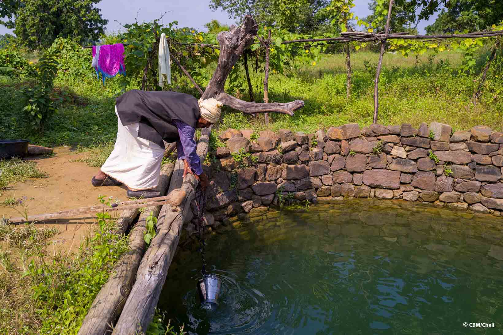 Un Indiano di mezza età con un handicap fisico chinato su un pozzo sta riempiendo d’acqua un secchio di metallo appeso a una catena. 
