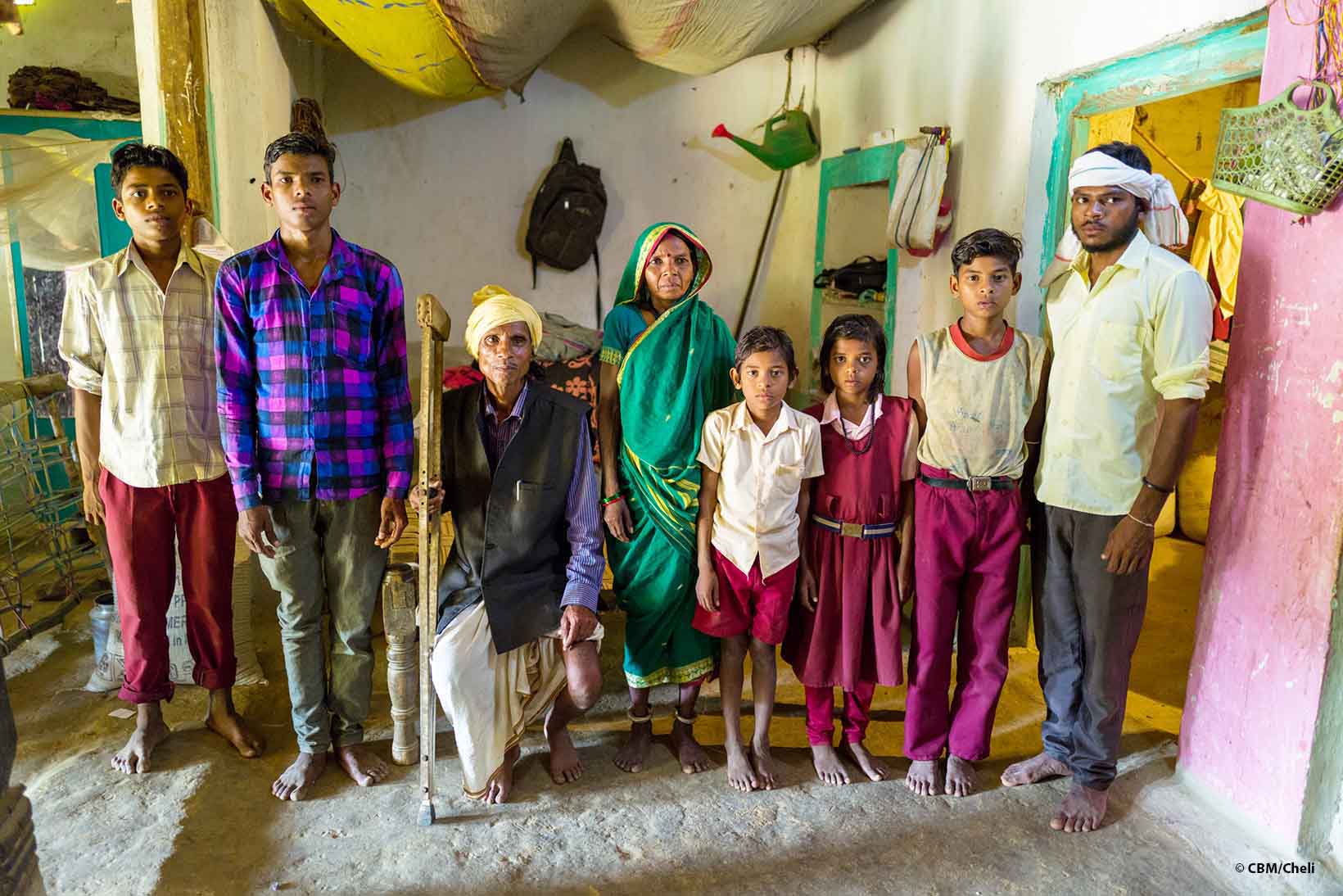 Ritratto di una famiglia indiana all’interno della loro casa. Il padre siede sul telaio di un letto e nella mano destra tiene una stampella di legno. In piedi alla sua sinistra c’è la moglie. Ai lati della coppia sono allineati in piedi i figli, cinque maschi e una femmina. 