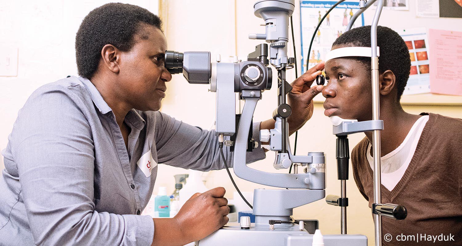 Teenagerin blickt ins Untersuchungsmikroskop. Die Ärztin hält ihr eine Linse vor ein Auge und schaut konzentriert ins Mikroskop.