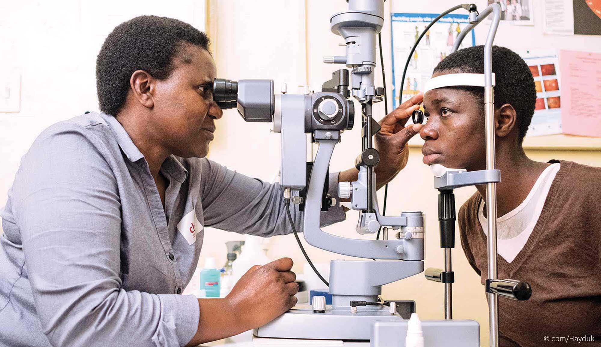 Teenagerin blickt ins Untersuchungsmikroskop. Die Ärztin hält ihr eine Linse vor ein Auge und schaut konzentriert ins Mikroskop.