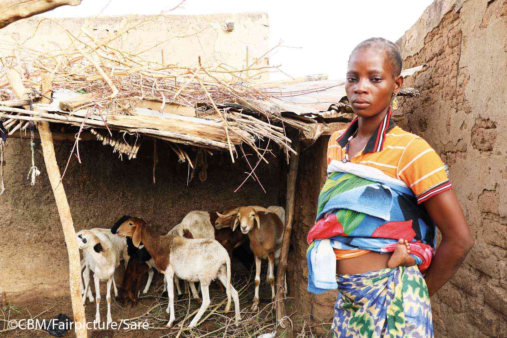 Miriam Sawadogo steht vor einem kleinen Stall. Im Stall stehen und fressen zwei Schafen und ein Schafsbock. 