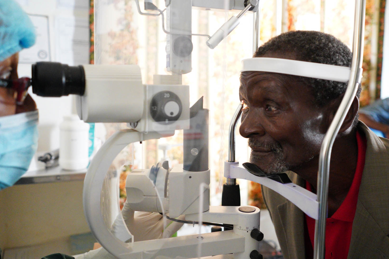 Un homme est assis devant un appareil d'examen et tient sa tête dans l'appareil. De l'autre côté, une médecin examine ses yeux.
