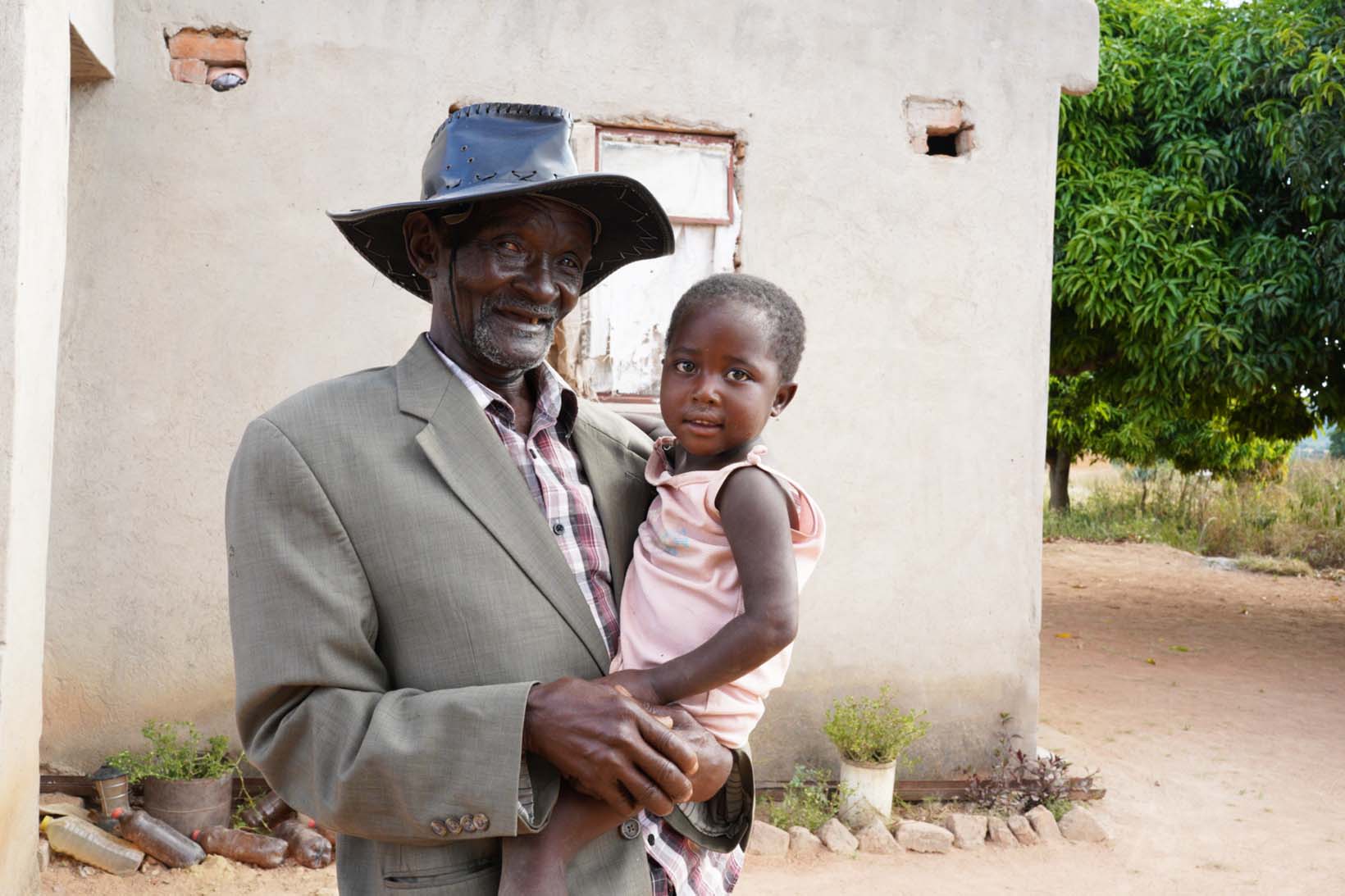 Paul steht vor seinem einfachen Haus und hat seine kleine Enkelin auf dem Arm. Beide lächeln.