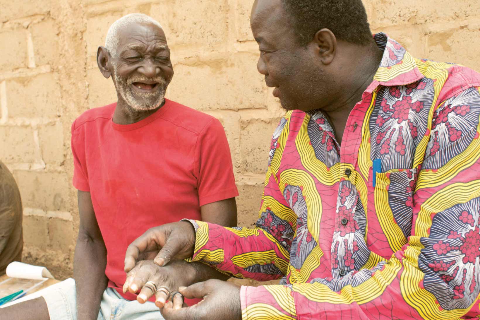 Ein Mann aus Burkina Faso sitzt auf einer Bank und hält die Hand von einem anderen Mann neben sich. Beide lächeln.