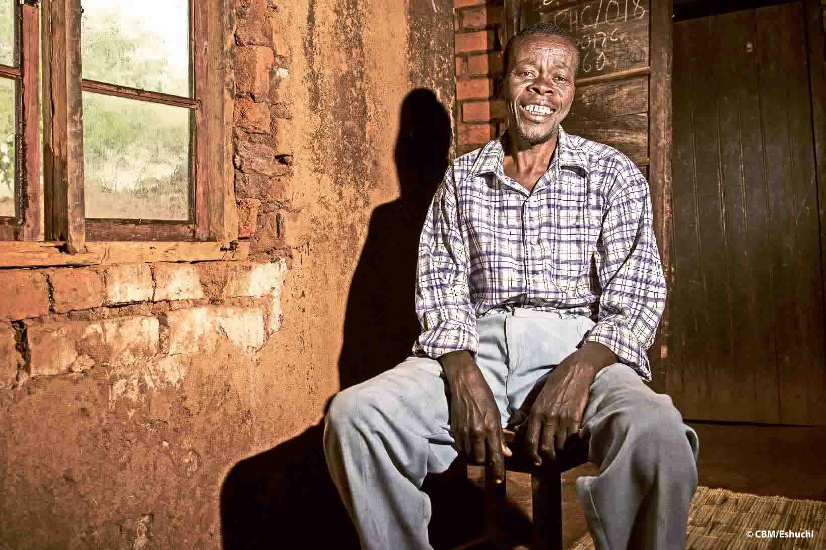 Un uomo del Malawi si siede su una sedia in una capanna di legno e sorride.