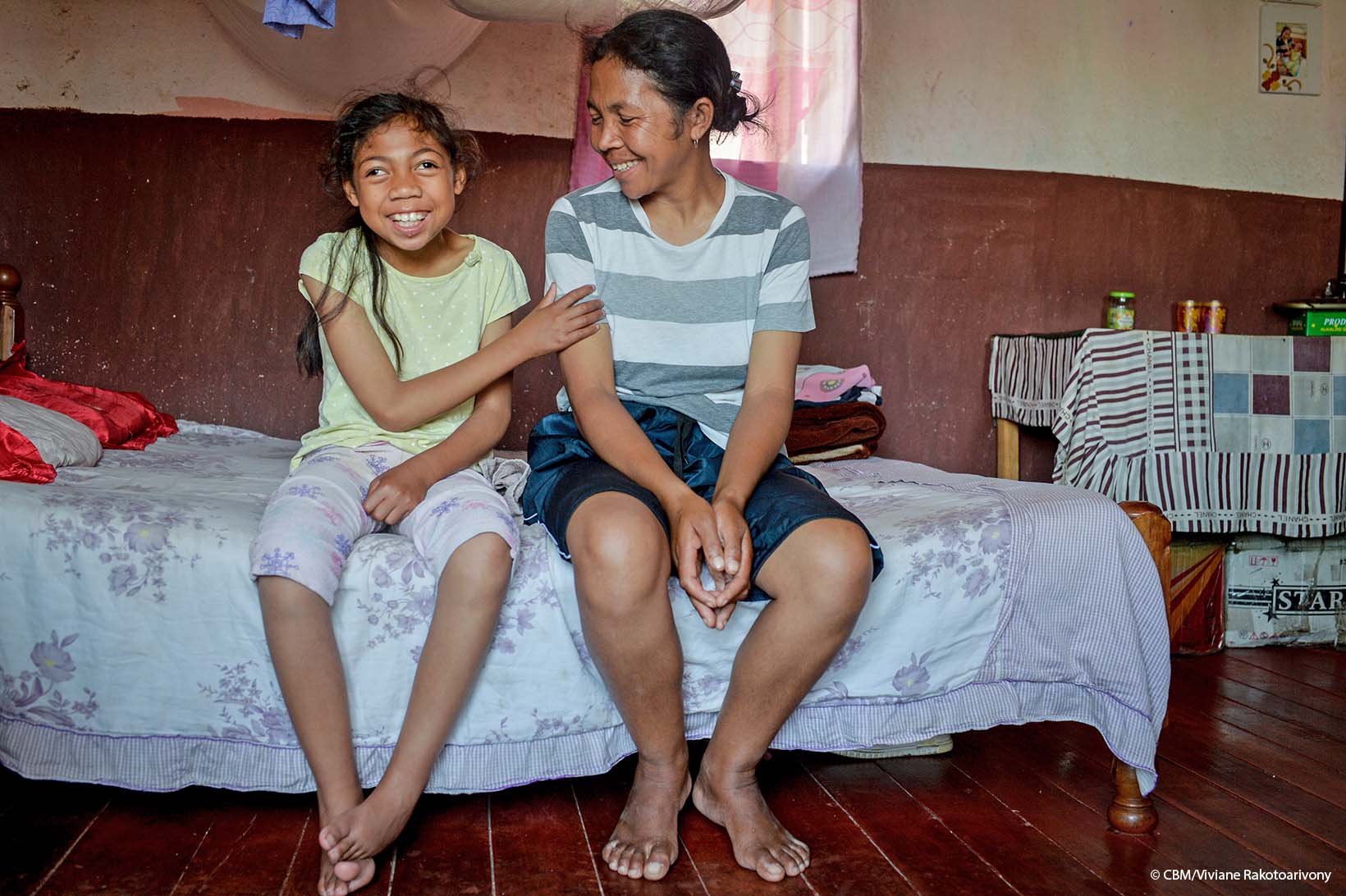 Ein Mädchen mit körperlicher und kognitiver Behinderung aus Madagaskar sitzt auf einem Bett und hält mir ihrer rechten Hand den linken Oberarm ihrer Mutter. Beide lächeln.