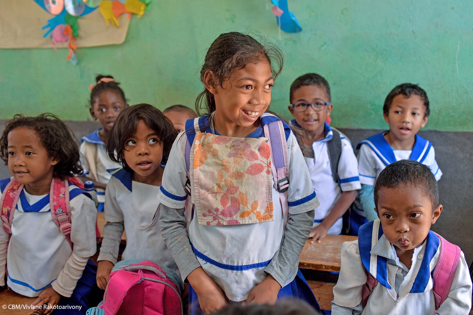 Ein Mädchen mit körperlicher und kognitiver Behinderung aus Madagaskar steht in einem Schulzimmer. Die Kinder um das Mädchen herum sitzen.