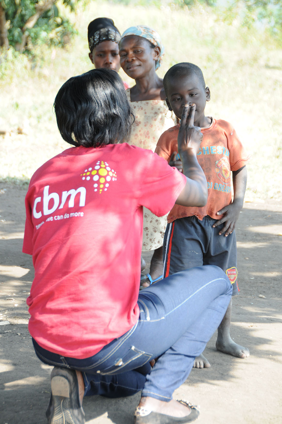 Une femme accroupie examine la vision d’un gamin en Ouganda au moyen d’exercices des doigts. A l’arrière plan, deux femmes l’observent.