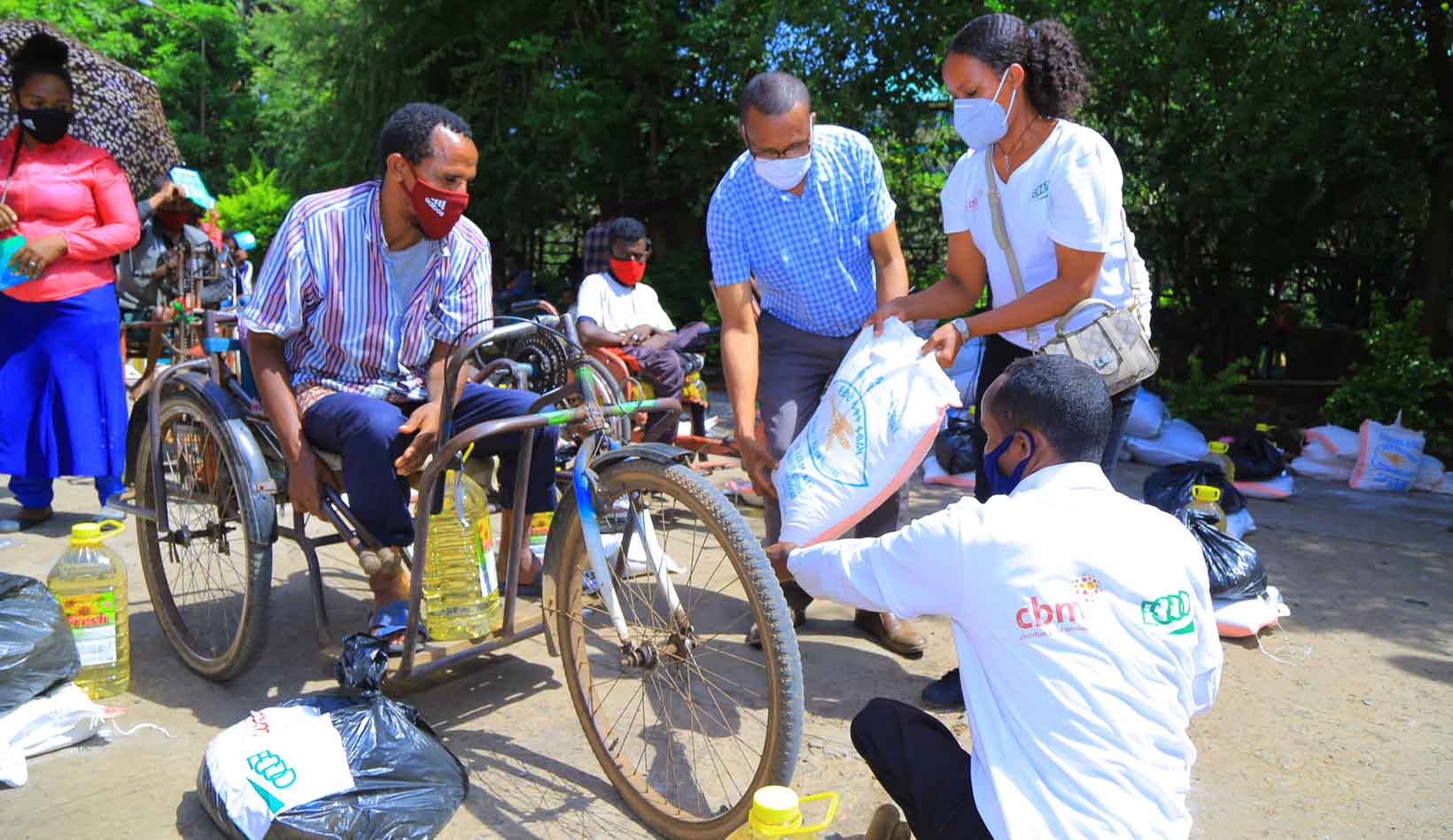 Ein Mann mit körperlicher Behinderung aus Äthiopien, der in einem Tricycle sitzt und eine Maske trägt, erhält von mehreren Personen Lebensmittel.
