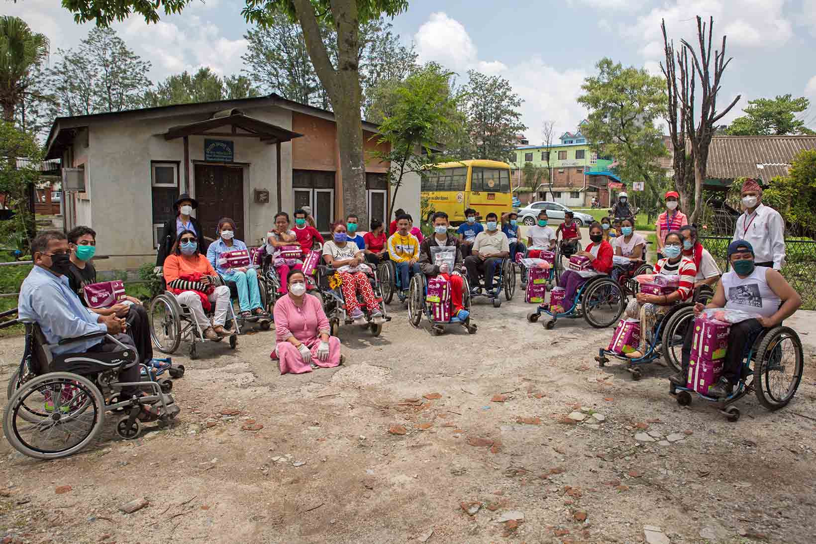 Diverse persone in sedia a rotelle in Nepal formano un semicerchio. In grembo o ai loro piedi hanno articoli per l’igiene. Al loro fianco si vedono alcuni assistenti. 