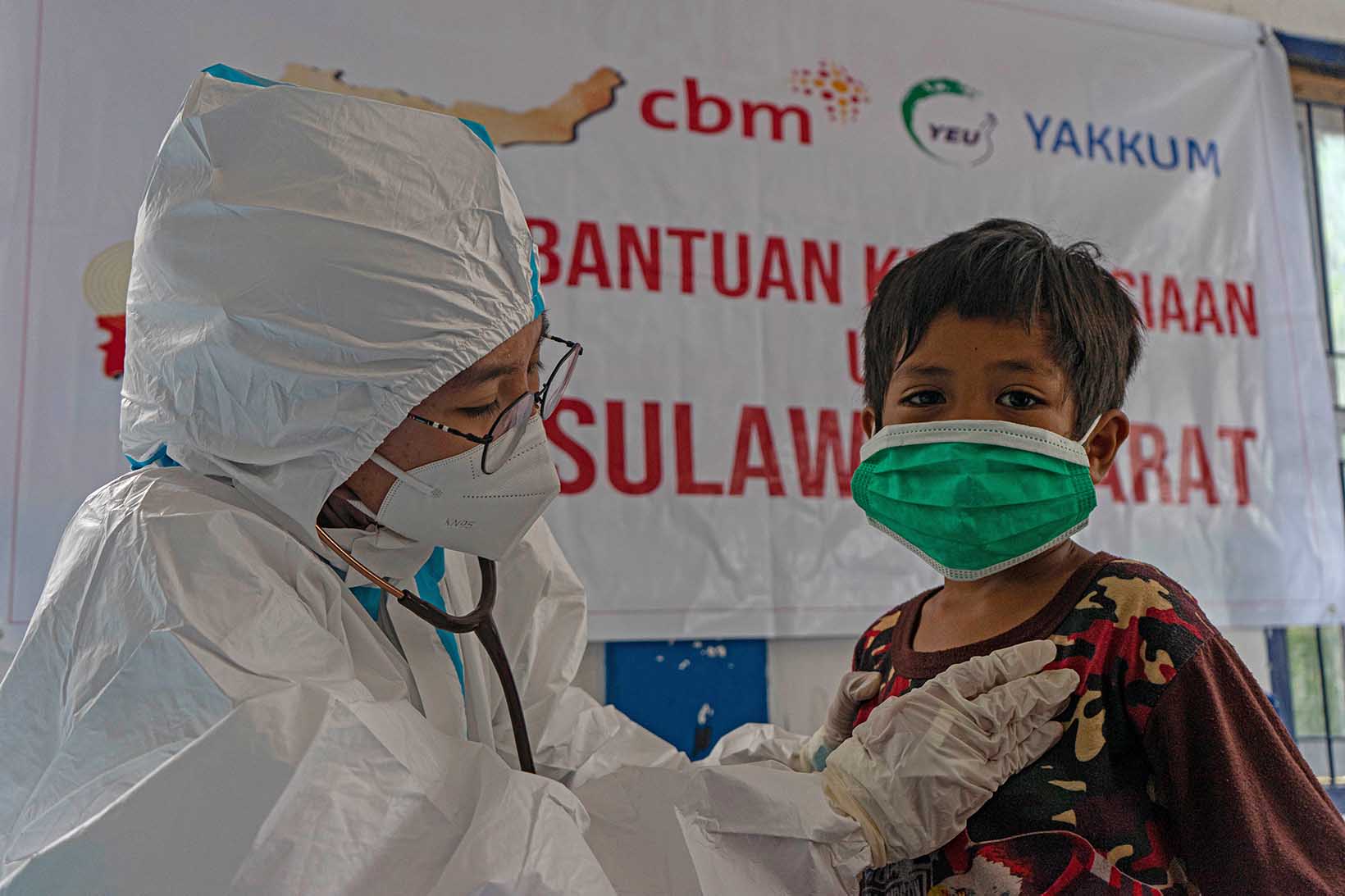 Dans une clinique mobile, une femme indonésienne en vêtement de protection anti-Covid ausculte au stéthoscope le cœur d’une fillette qui porte un masque de protection.