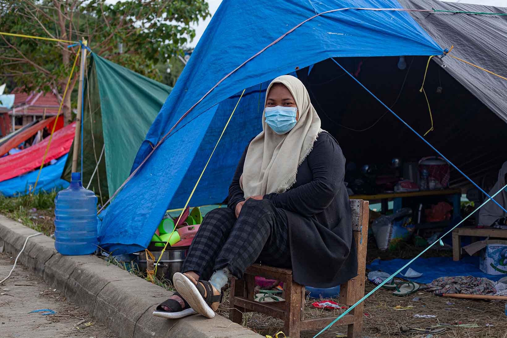 Una donna di Sulawesi con una protesi alla gamba sinistra è seduta su una sedia davanti a una tenda per sfollati. Indossa una mascherina.