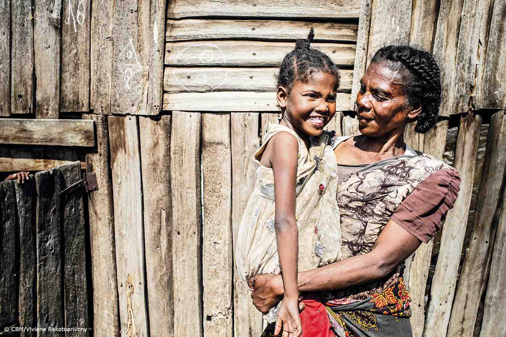 Une femme d'âge moyen aux cheveux soigneusement tressés regarde le visage amusé d'une petite fille dans ses bras. Elles se tiennent toutes deux devant une maisonnette en planches dans le sud de Madagascar.