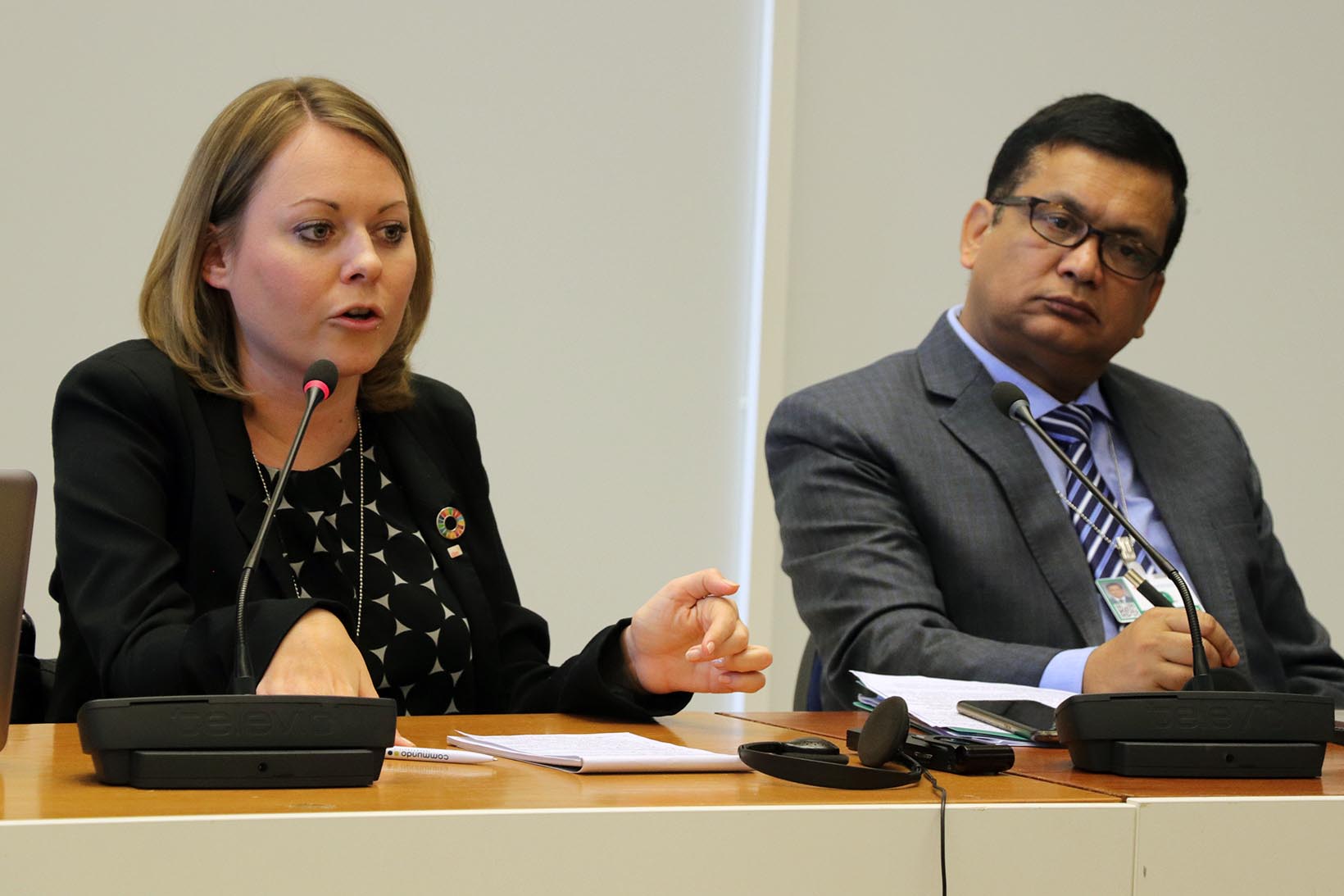 Eine Frau spricht an einer Konferenz, neben ihr sitzt ein Mann.