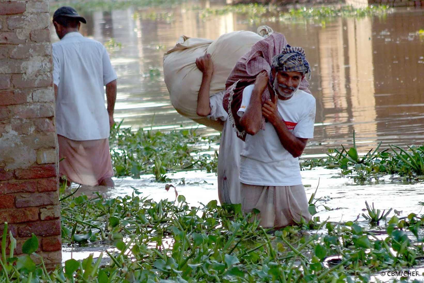 Drei Personen aus Pakistan gehen nach einer Überschwemmung im knietiefen Wasser und tragen Säcke über ihren Schultern.