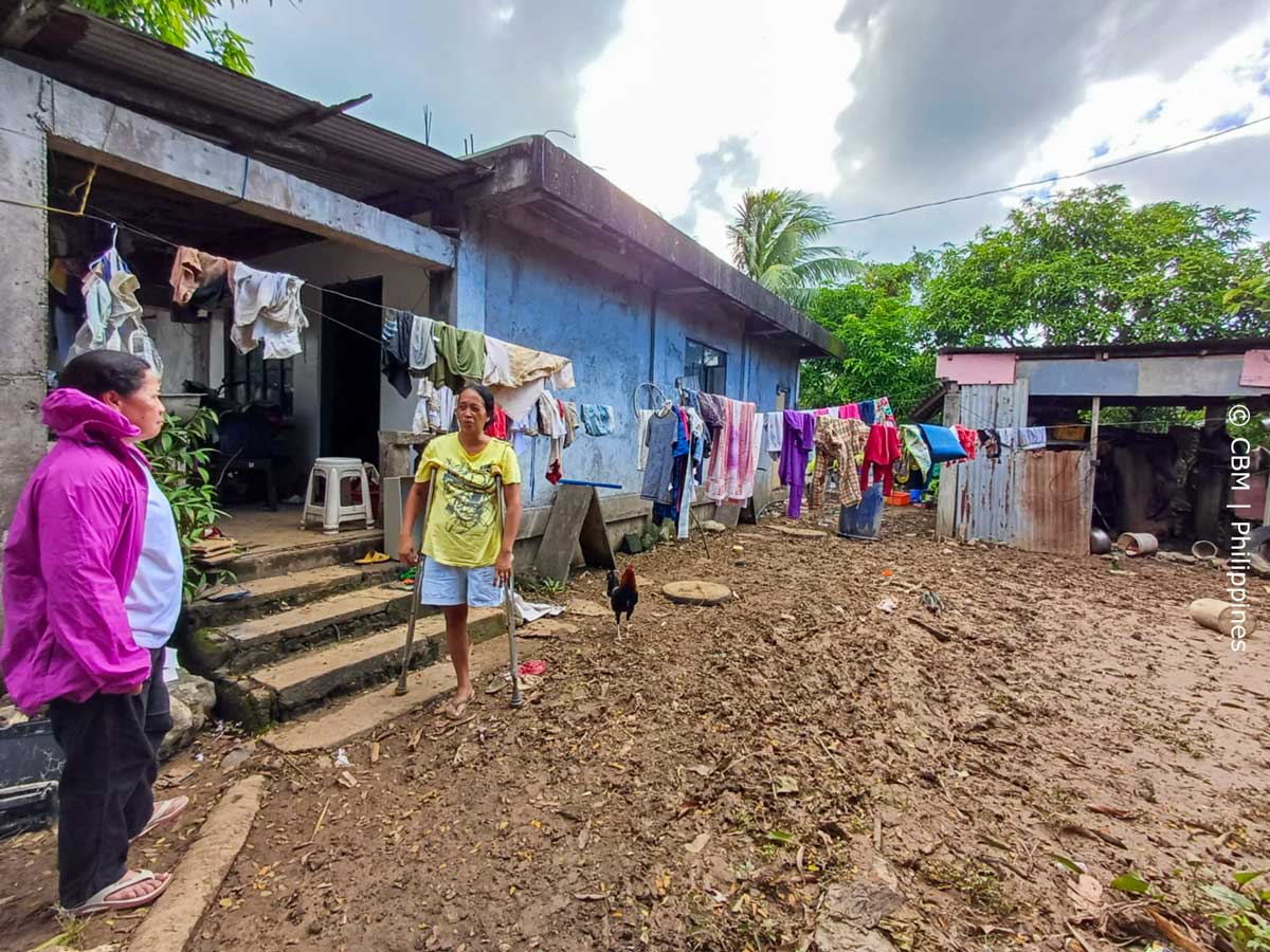 Ein überschwemmter schwammiger Hof mit zwei Häusern. Mittendrin eine Frau mit Krücken. 
