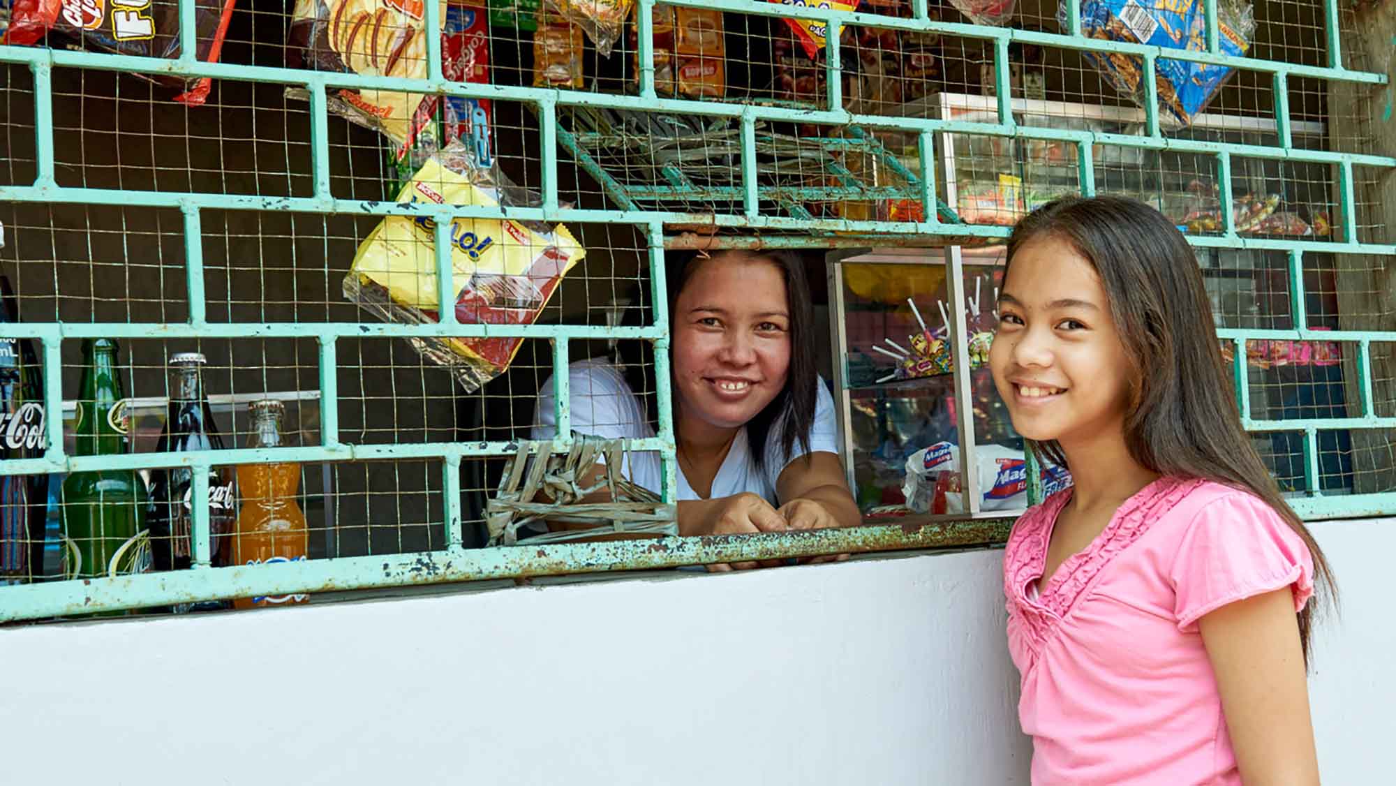 Durch die Verkaufsluke eines Kiosks lächelt eine Frau aus den Philippinen, die eine psychosoziale Behinderung hat, in die Kamera. Vor dem Kiosk steht ihre Tochter, die ebenfalls lächelt.