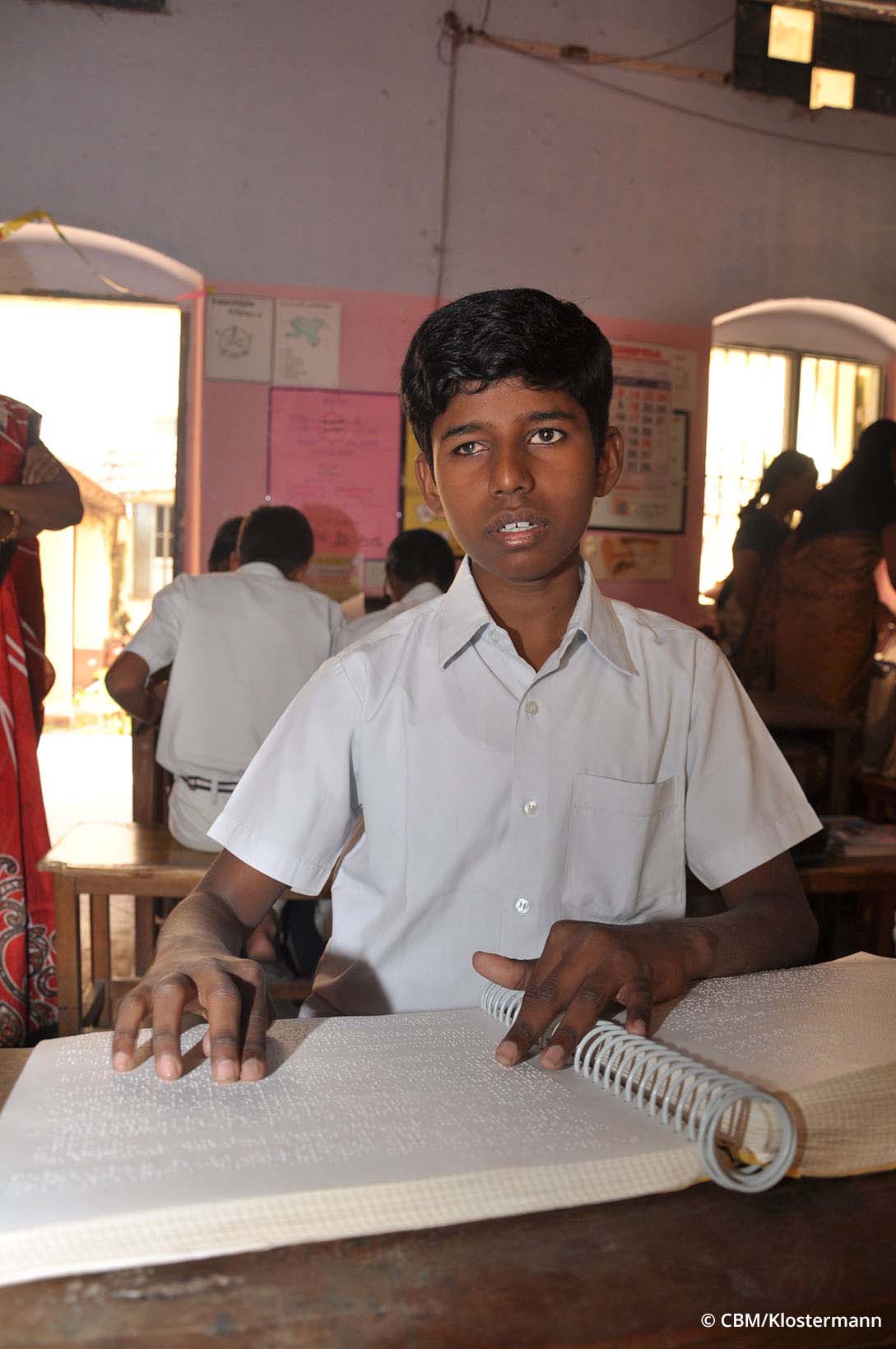 Ein Jugendlicher aus Indien liest in einem Schulzimmer aus einem Buch mit Brailleschrift.