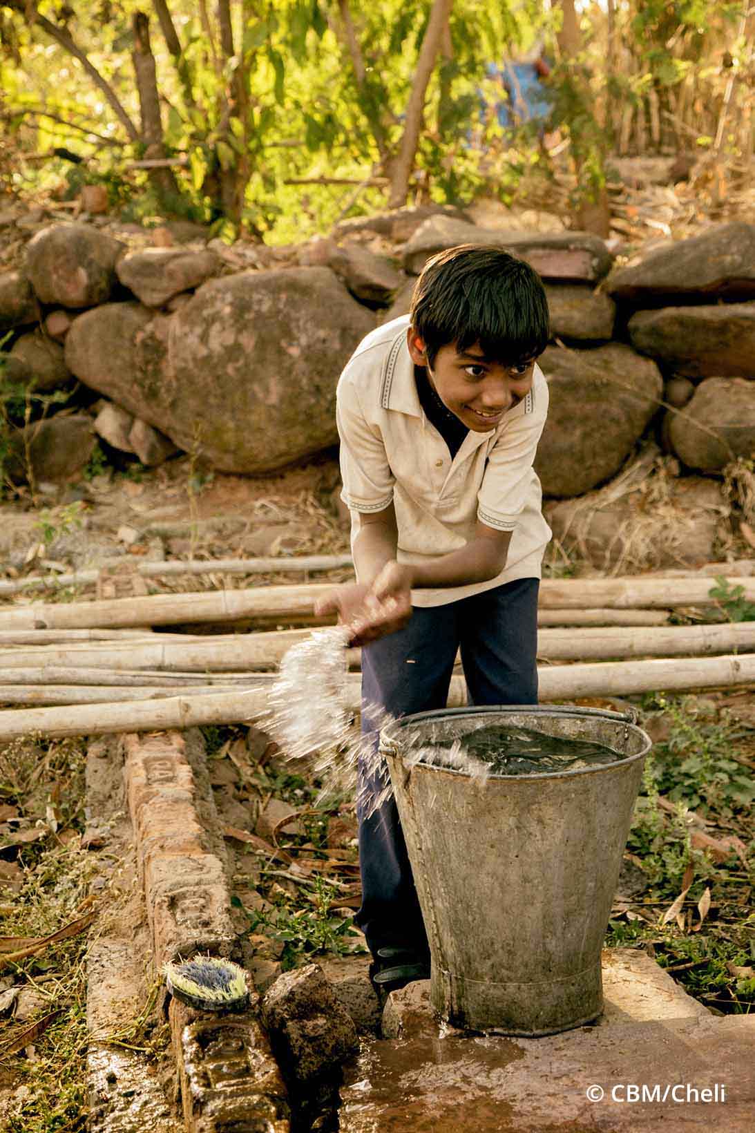 En Inde, un garçon puise de l'eau avec ses mains dans un seau et sourit.