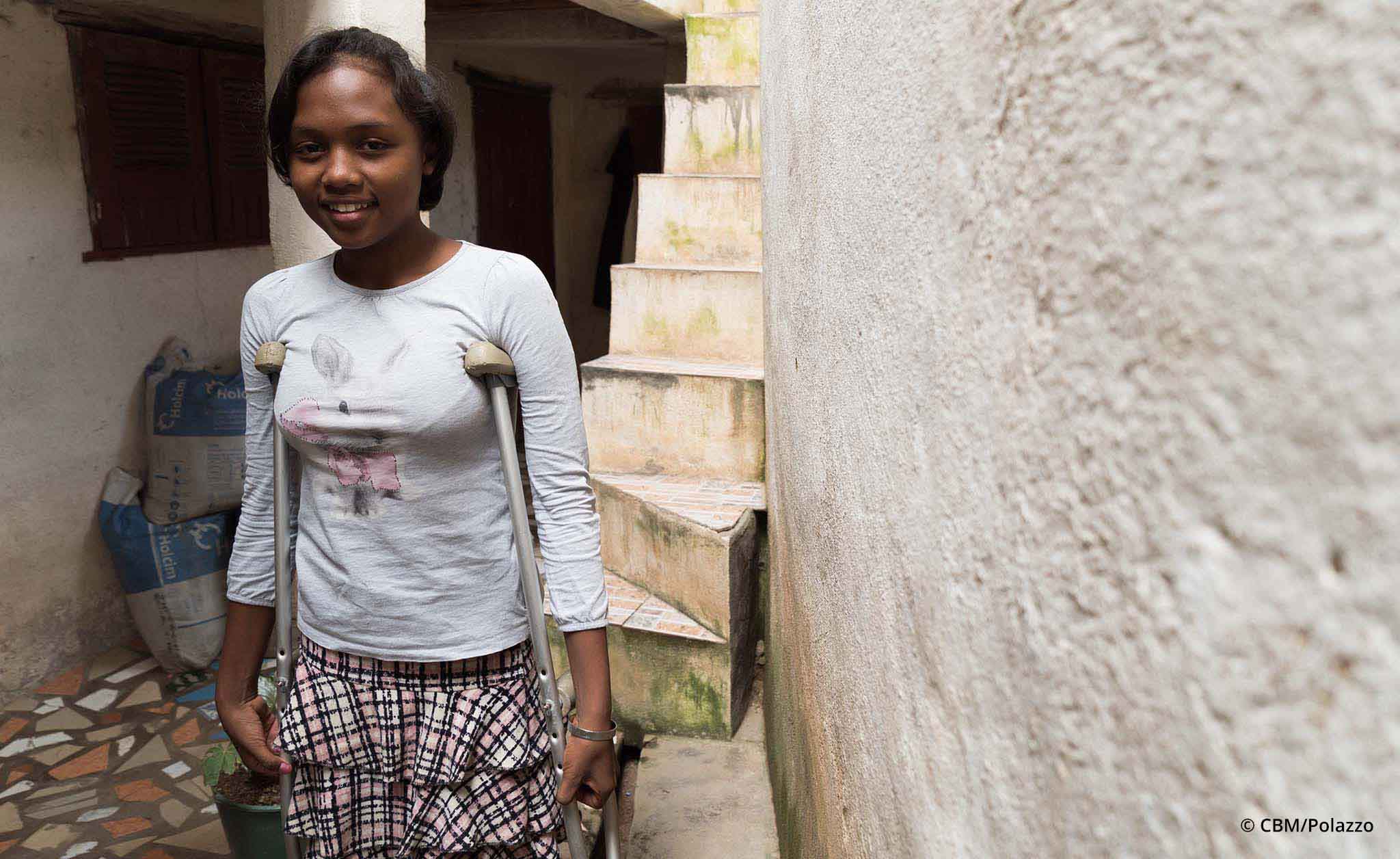 Eine junge Frau aus Madagaskar, die eine körperliche Behinderung hat, steht an Krücken an einer Hauswand.