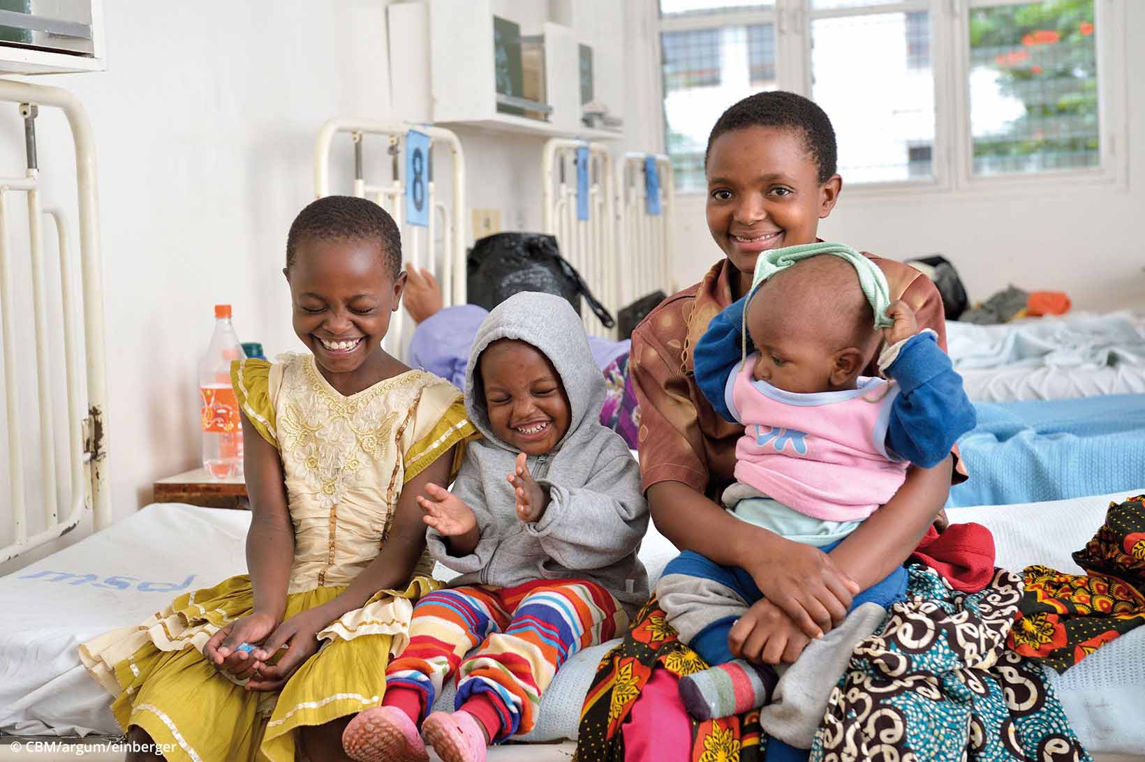 Eine Mutter aus Tansania sitzt mit ihren drei Kinder lächelnd auf einem Spitalbett.