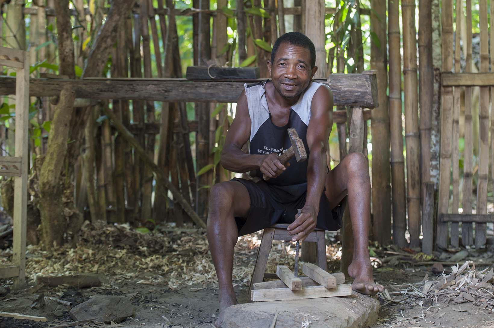 Ein freundlich blickender Mann aus Madagaskar bearbeitet mit Hammer und Stechbeitel zukünftige Stuhlbeine. Er sitzt auf einem Hocker vor seiner Werkbank im Freien. 